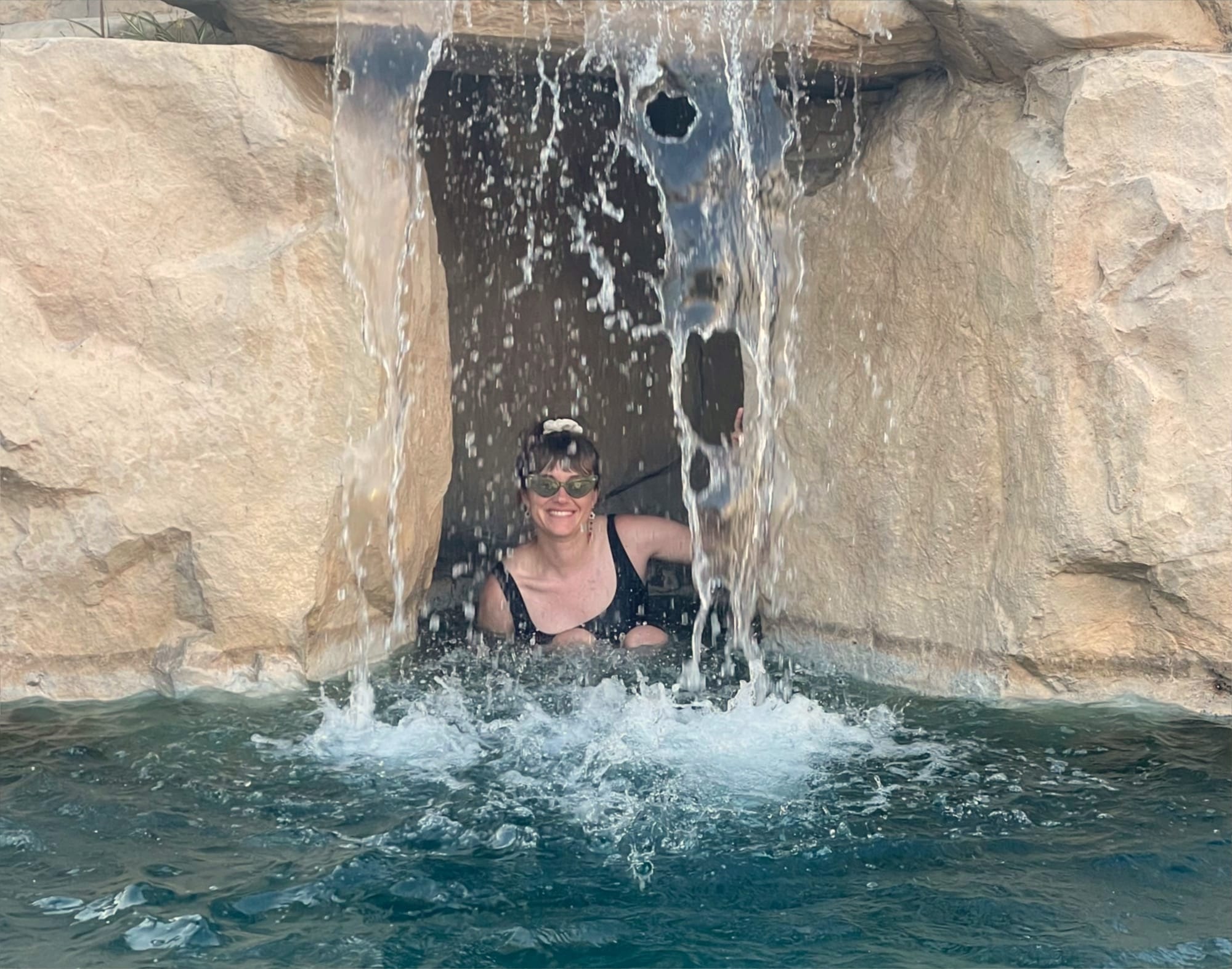 A woman in a pool between large beige rocks and beneath a waterfall.