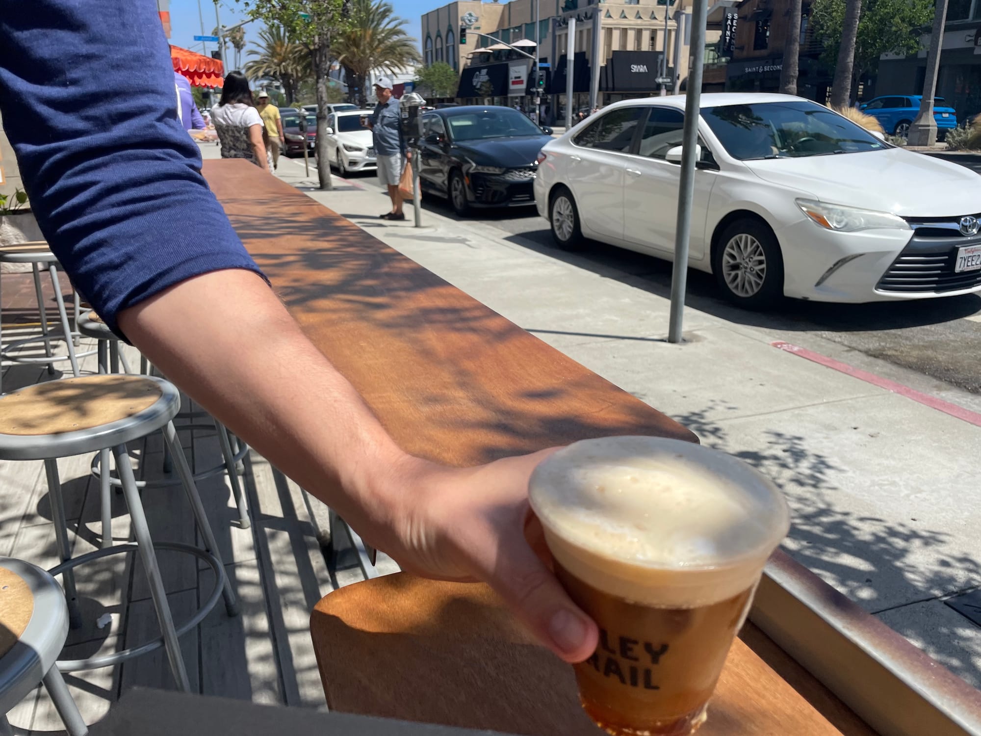 A hand holding a brown frothy beverage outside near a sidewalk lined with parked cars.