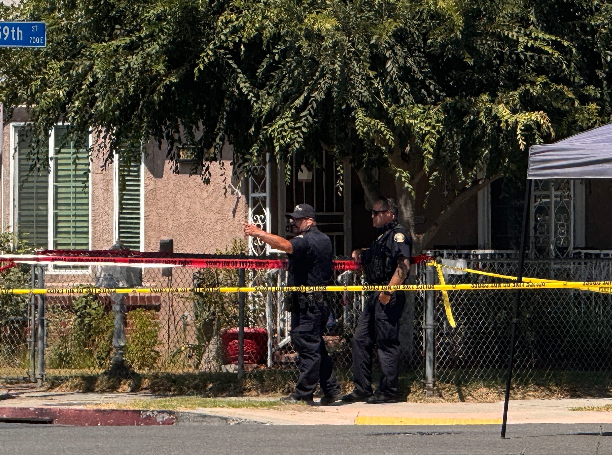 Two police officers in black uniforms stand in front of a tree and a house.