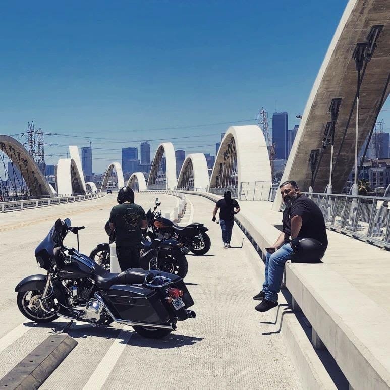 Two men stand and one man sits near three motorcycles on a bridge.