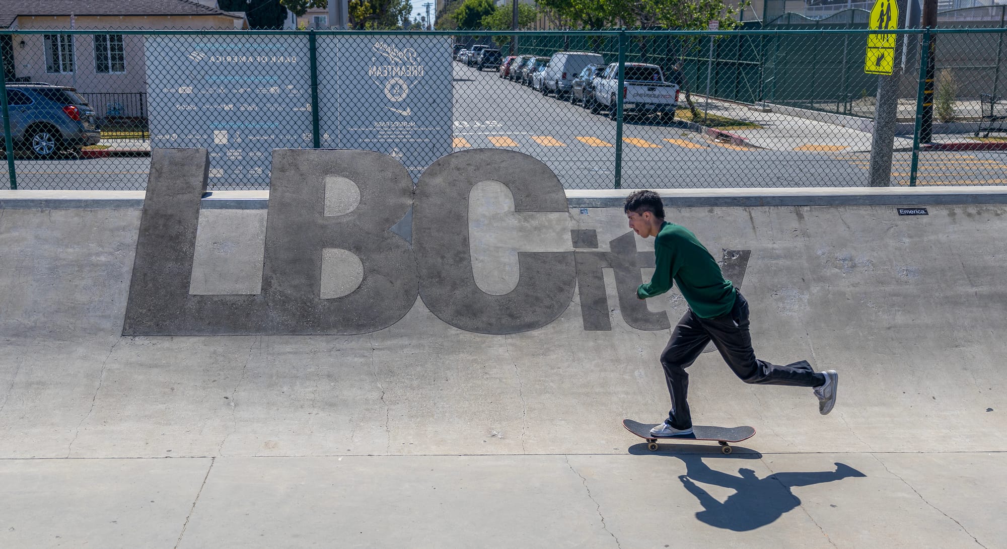 A skateboarder wearing a dark green long-sleeve shirt and dark pants rides past letters that spell L B City.