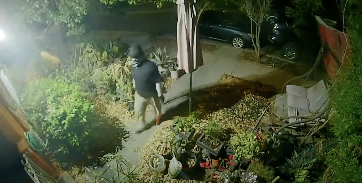 Surveillance image of a person wearing a dark shirt and light pants standing in a front yard at night.