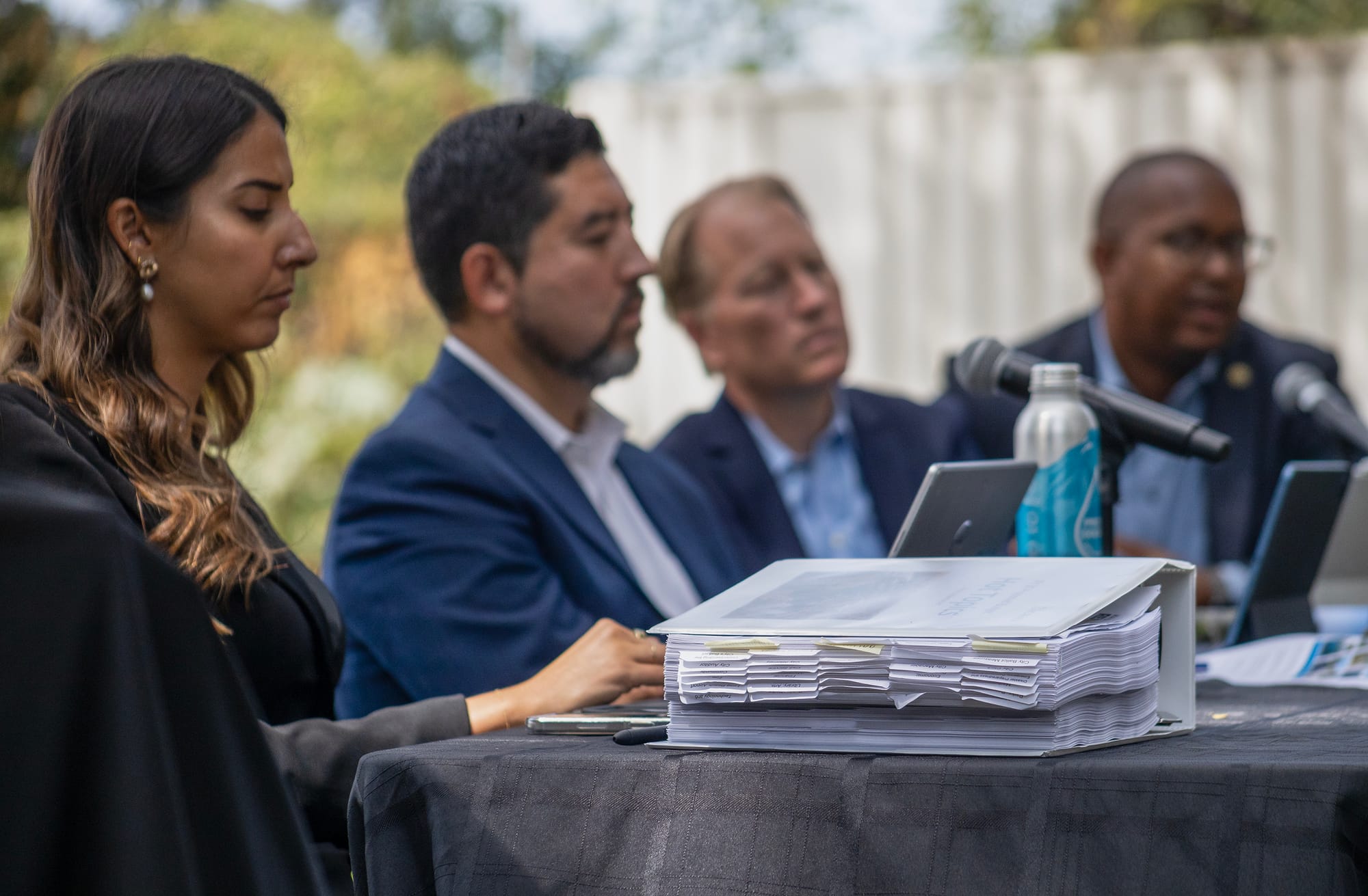 One woman and three men sit at a table with two laptop computers and a thick binder of papers.