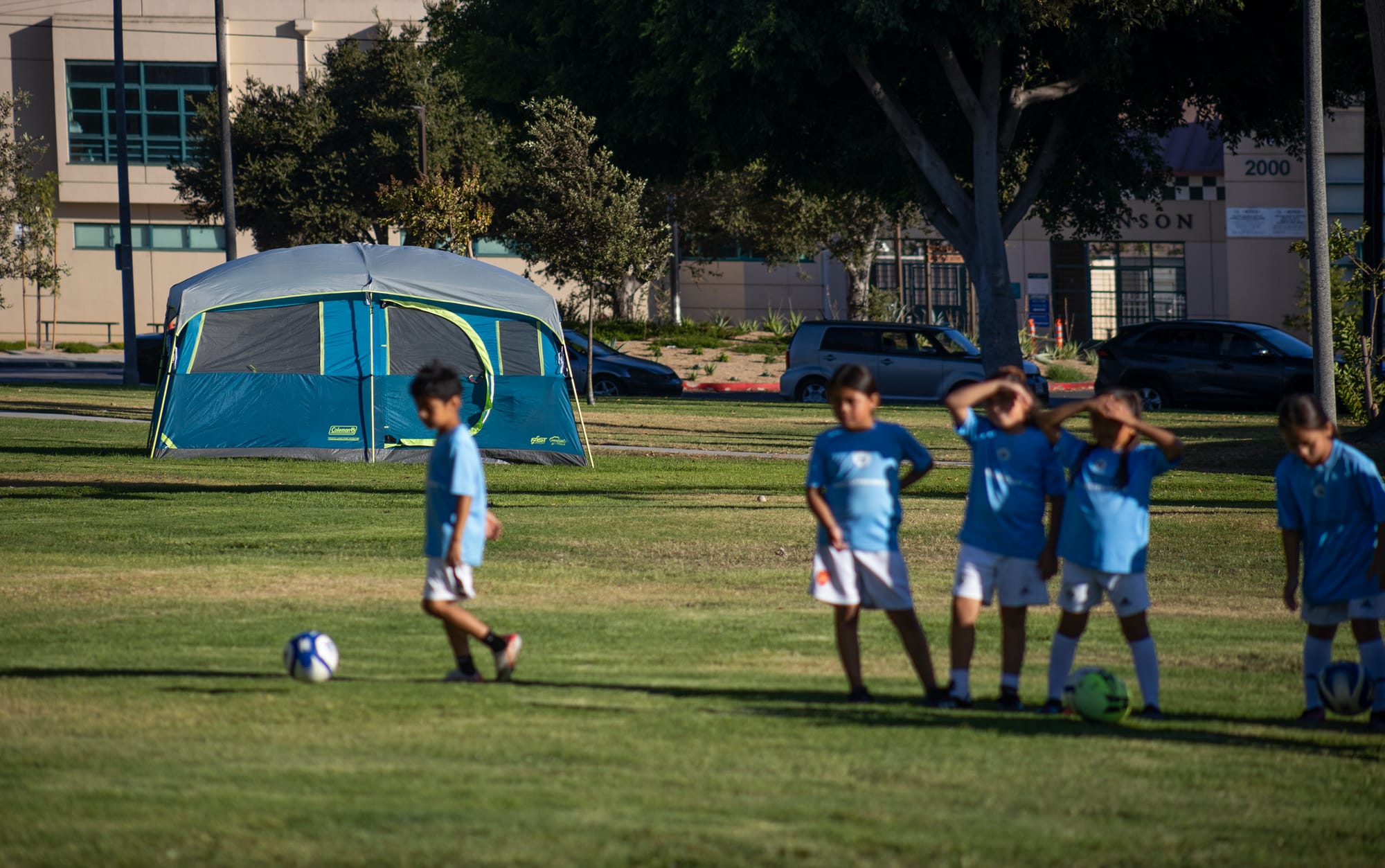 Five young children in blue and white jerseys play soccer at a park with a tent behind them.