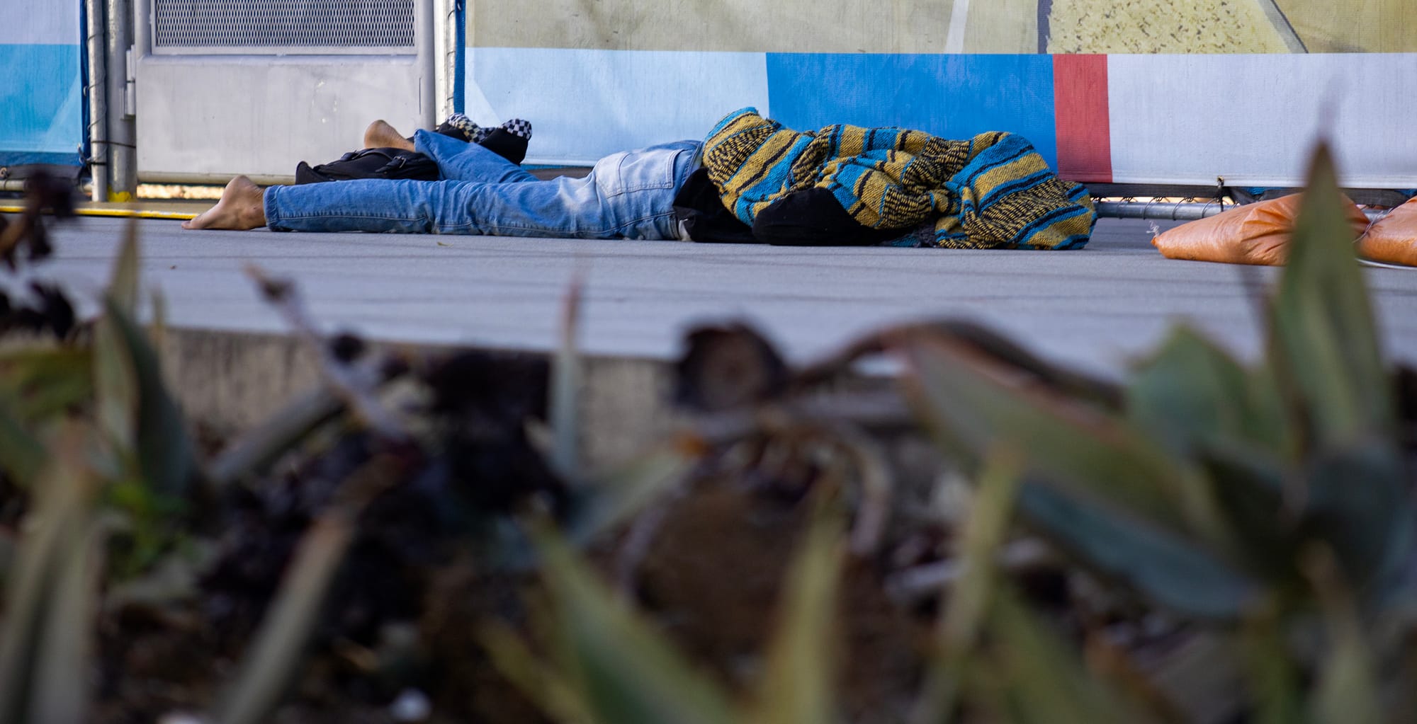 A bare-foot person covered with a yellow and blue blanket and wearing jeans lays on concrete.