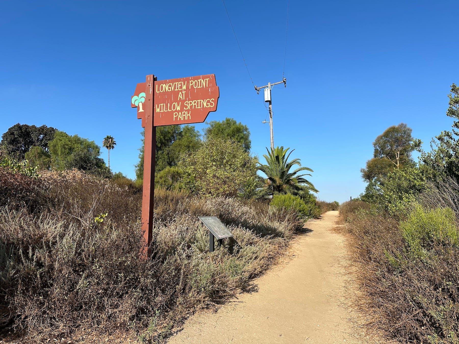 Brown sign saying "Longview Point at Willow Springs Park" next to a dirt trail through green brush.