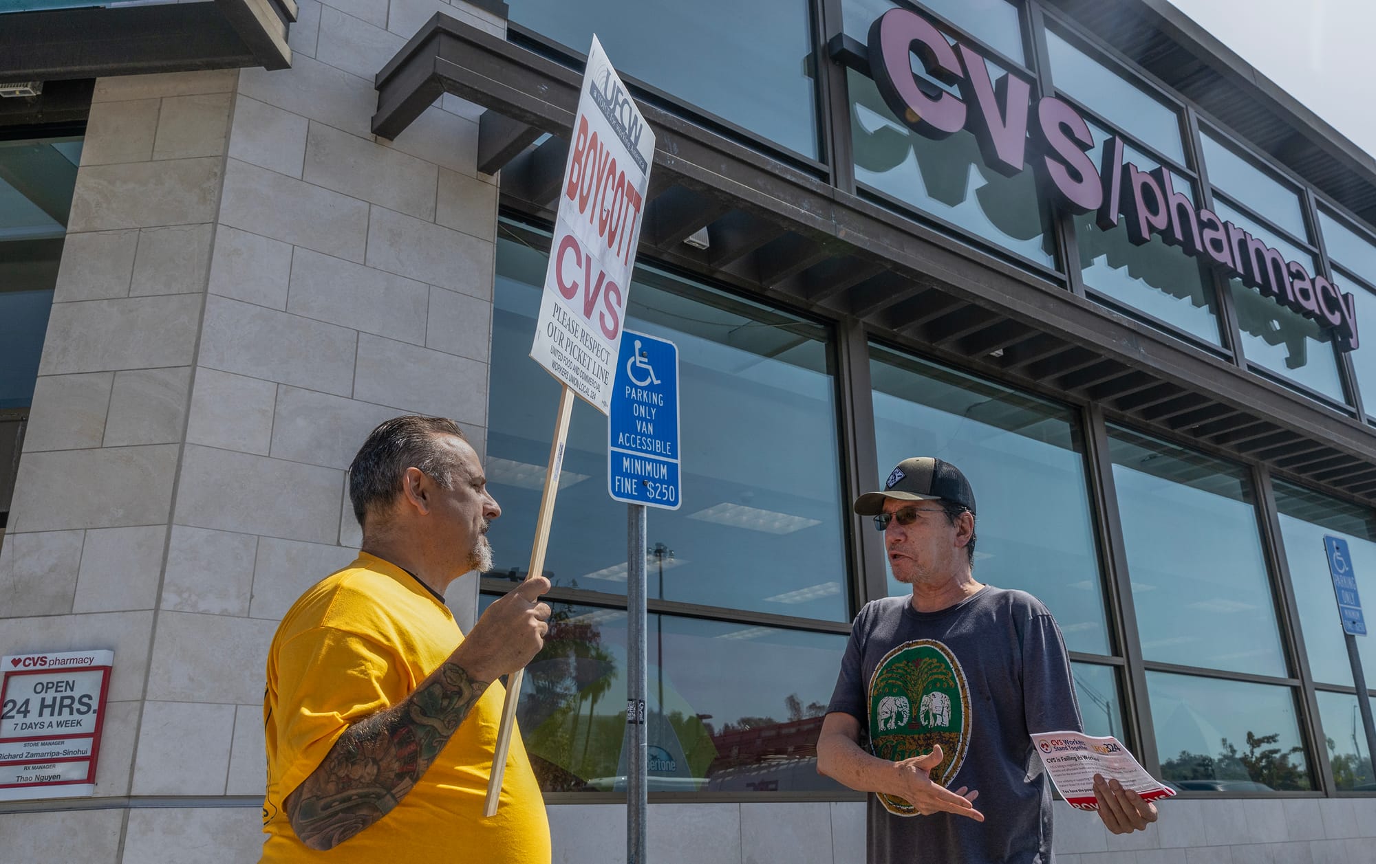 A man wearing a yellow shirt and holding a sign that says "boycott CVS" speaks to another man wearing a hat and sunglasses.