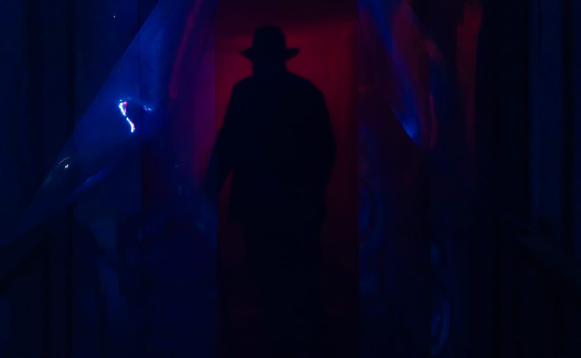 A man in a fedora stands in the shadows.