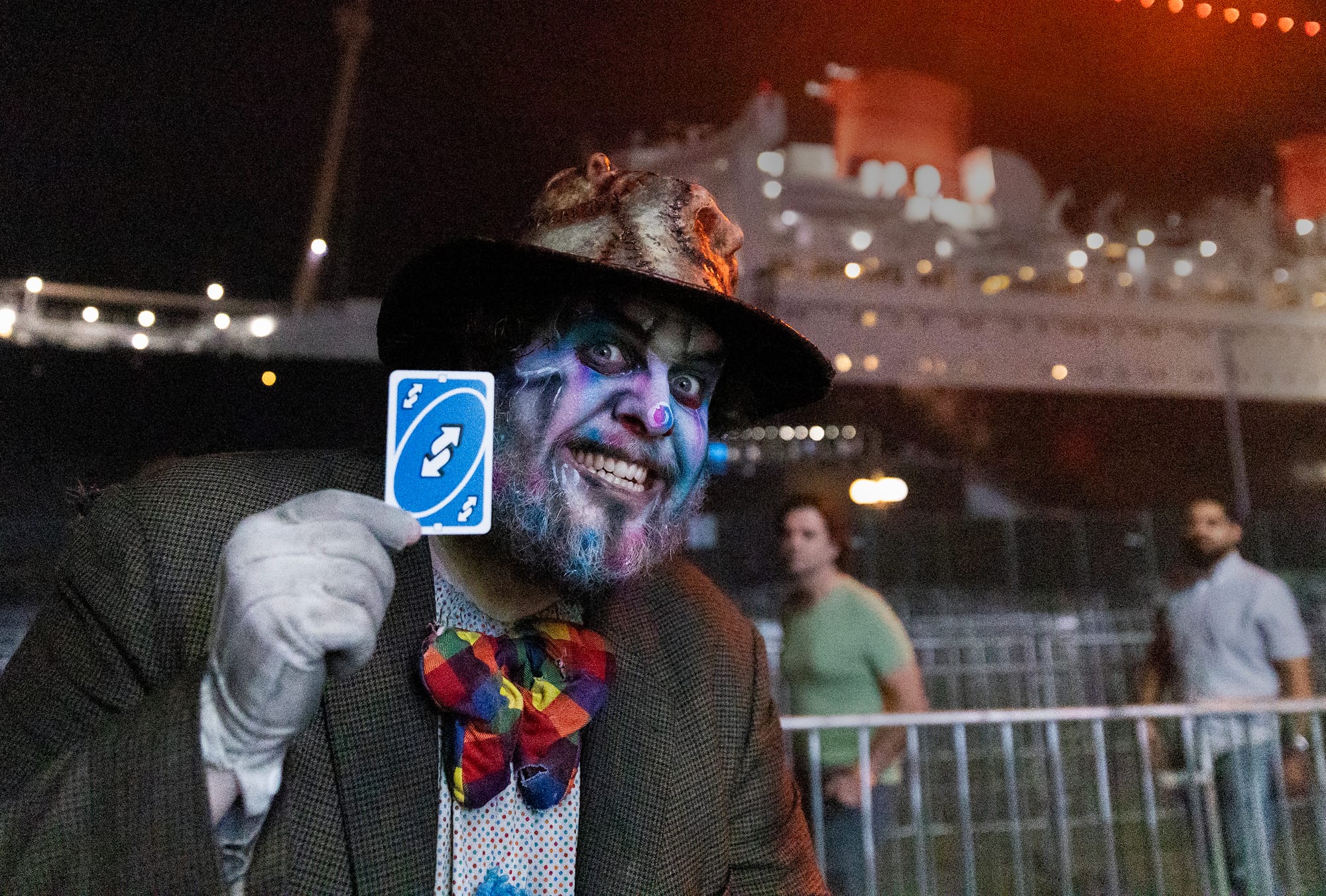 A man in a hat and heavy makeup holds up a playing card in front of the Queen Mary ocean liner.