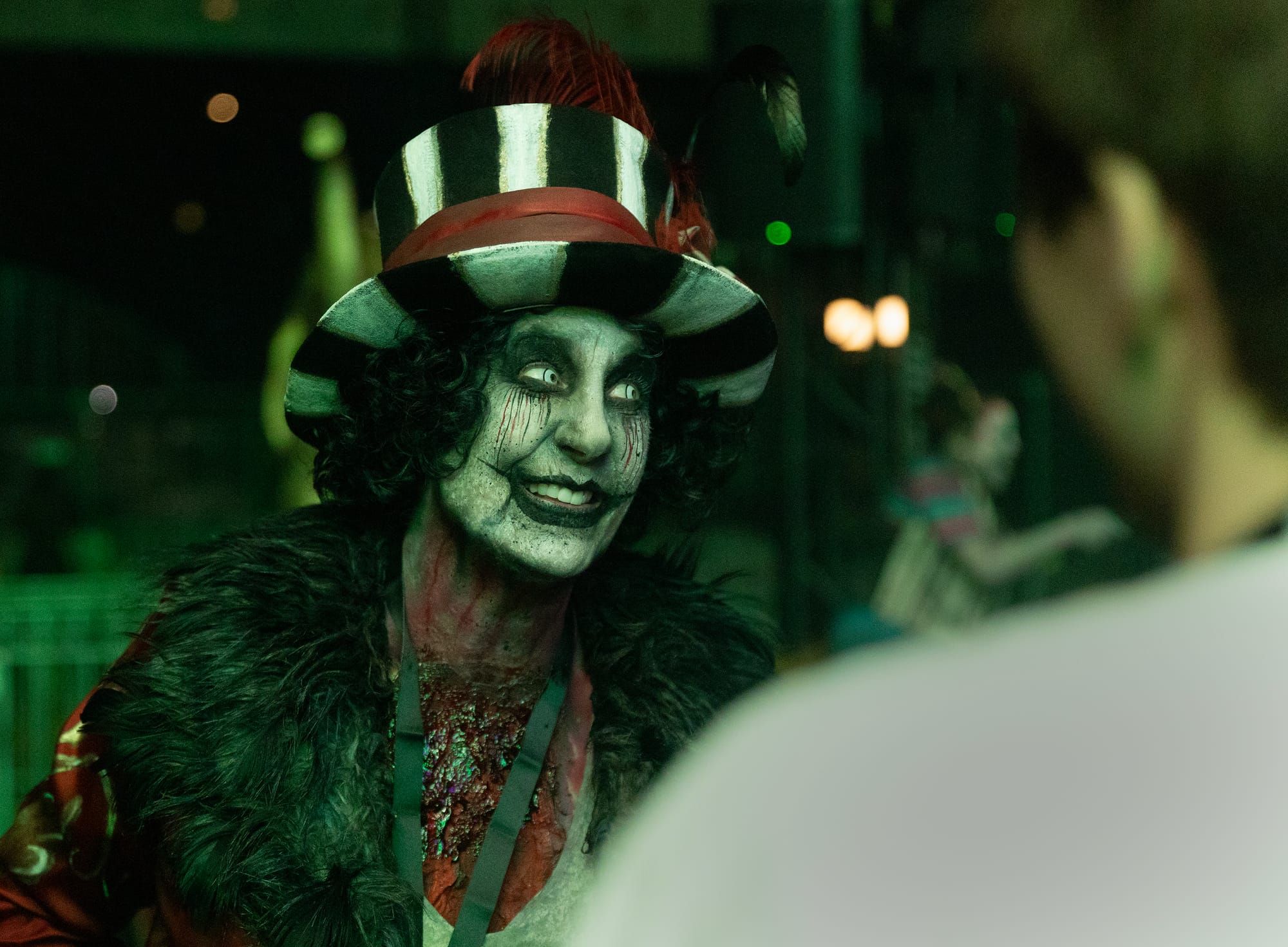 A person in heavy face paint wears a large ringmaster hat.