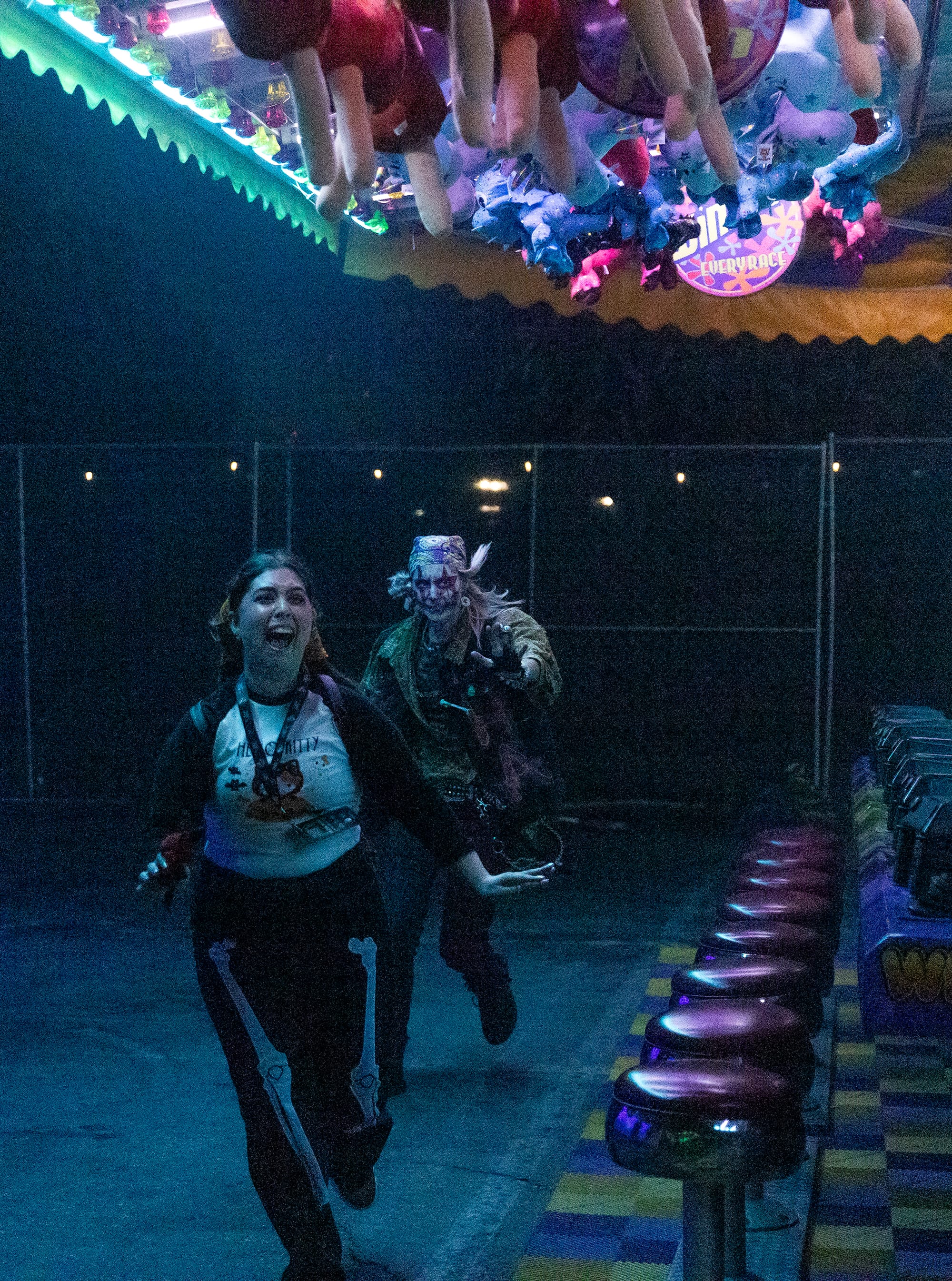 A person in scary makeup chases a woman next to a line of stools.