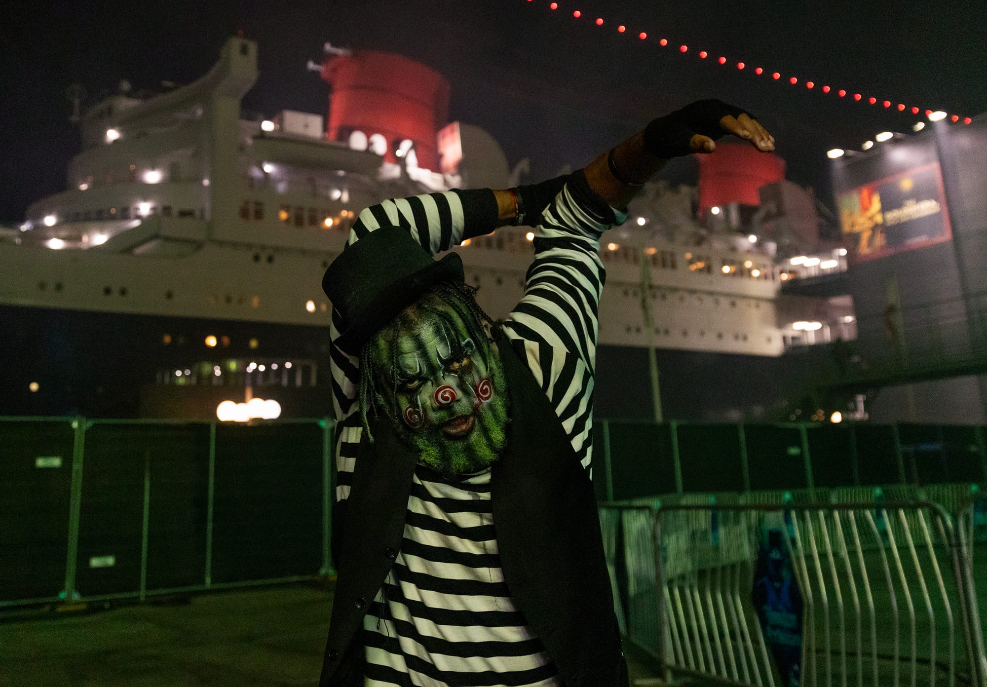 A man in heavy horror makeup contorts his arms in front of the Queen Mary luxury liner.