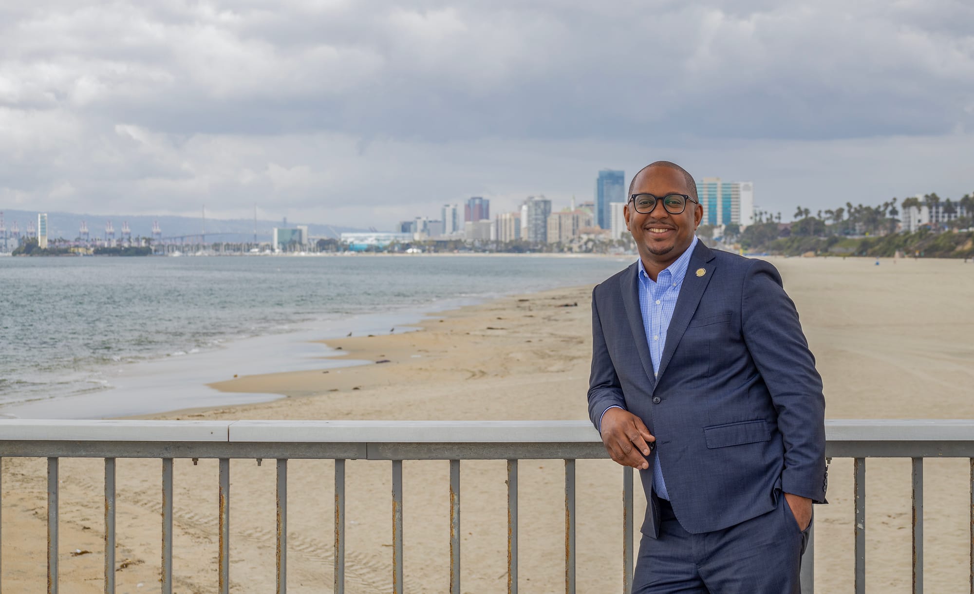 A man in a navy blue suit leans on a rail with a beach and city skyline in the background.