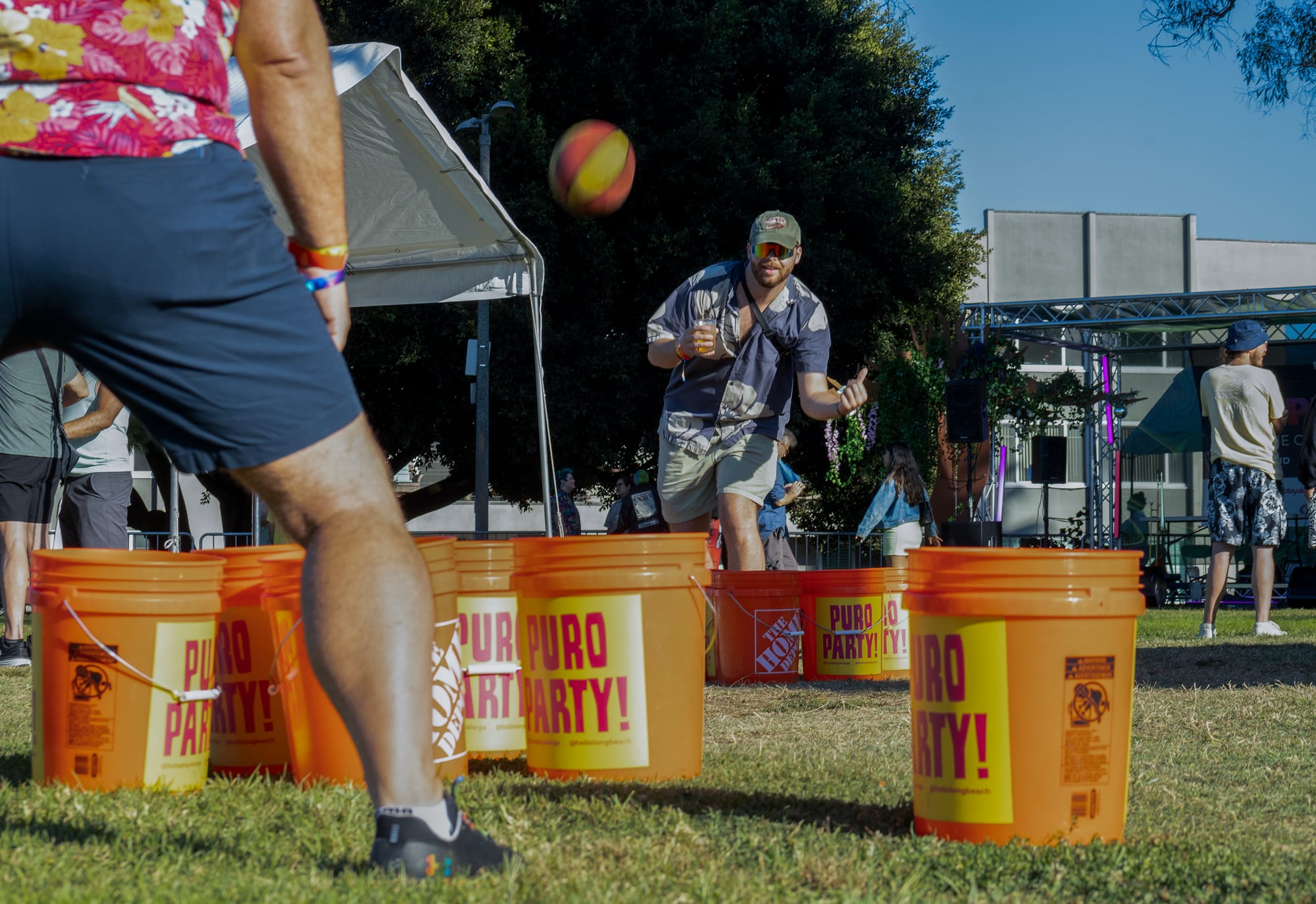 A man wearing a hat and sunglasses, and holding a drink, throws a ball.