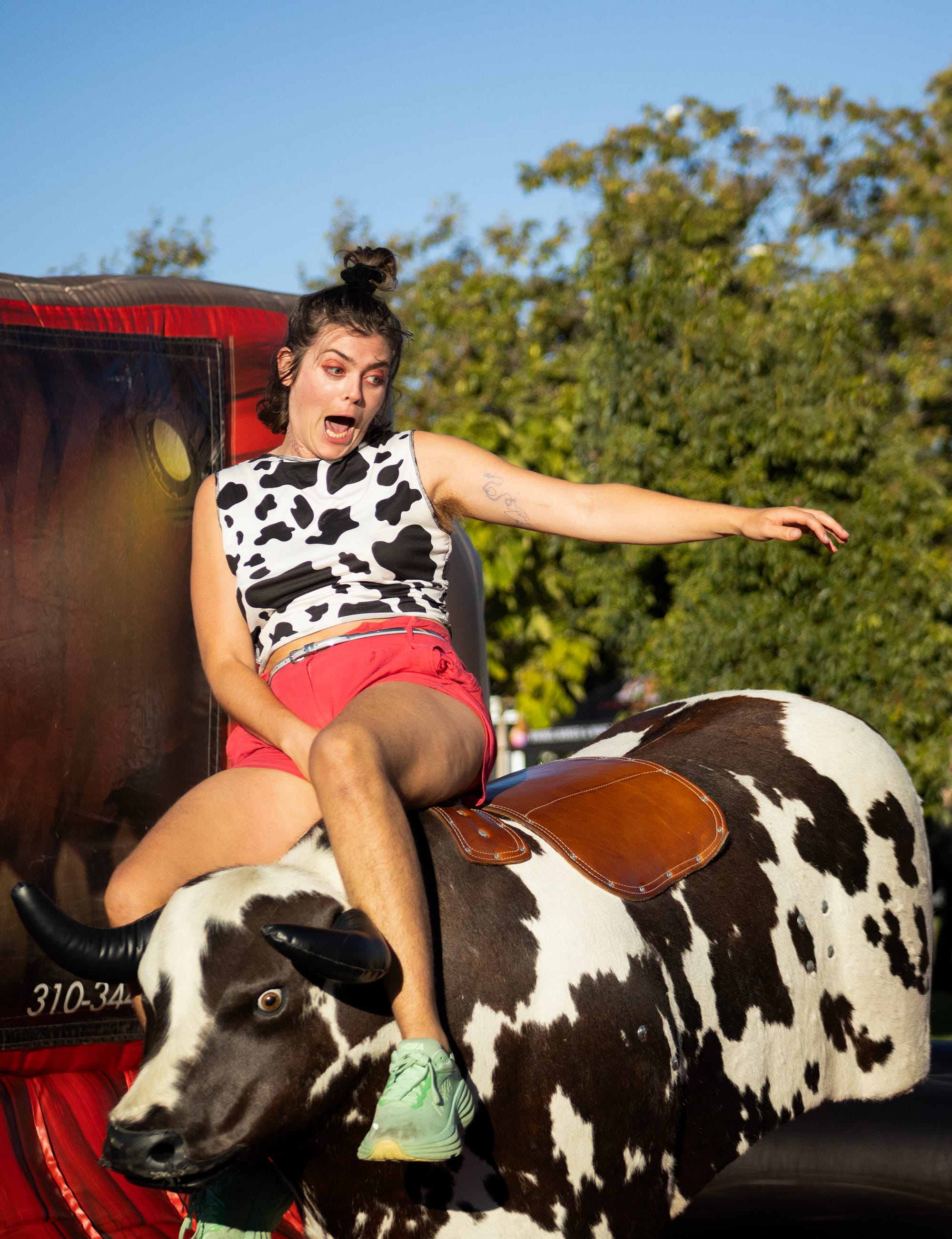 A woman wearing a cow-print shirt rides a mechanical bull.