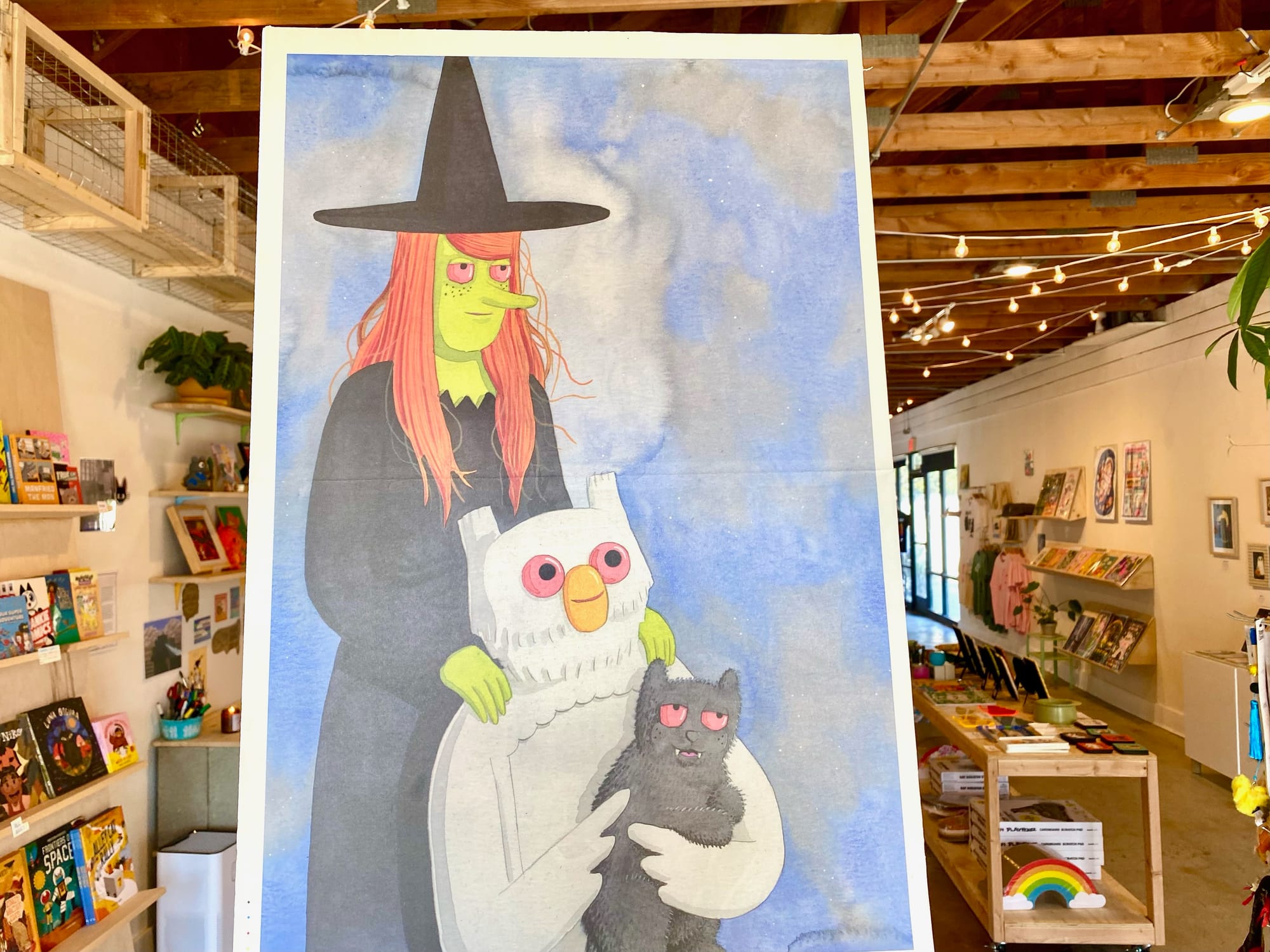 A painting of a green-skinned witch resting her hands on a white owl that's holding a black cat.