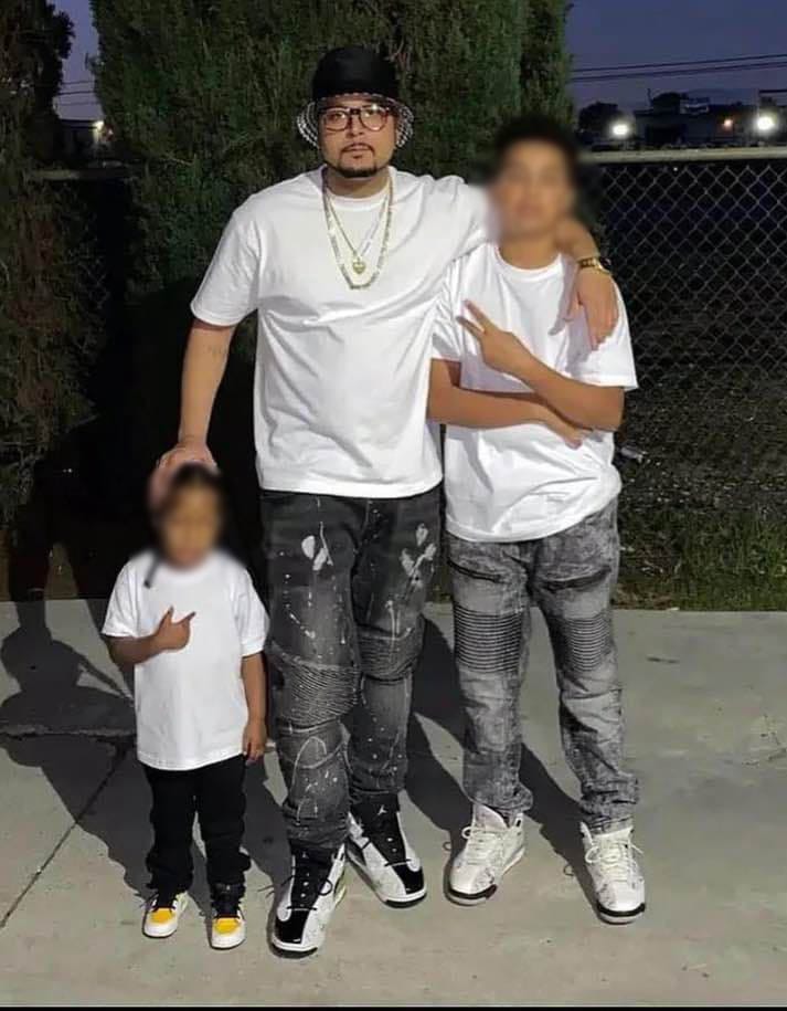 A man wearing a bucket hat, glasses, a white shirt and black jeans stands with two young boys.