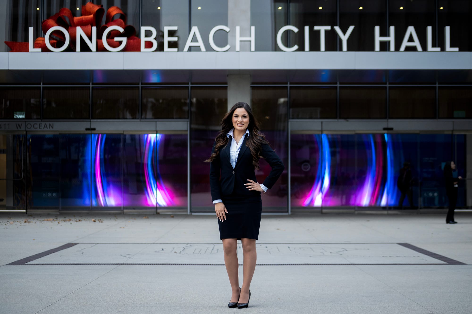 A young woman in dark business attire stands in front of a building marked "Long Beach City Hall."