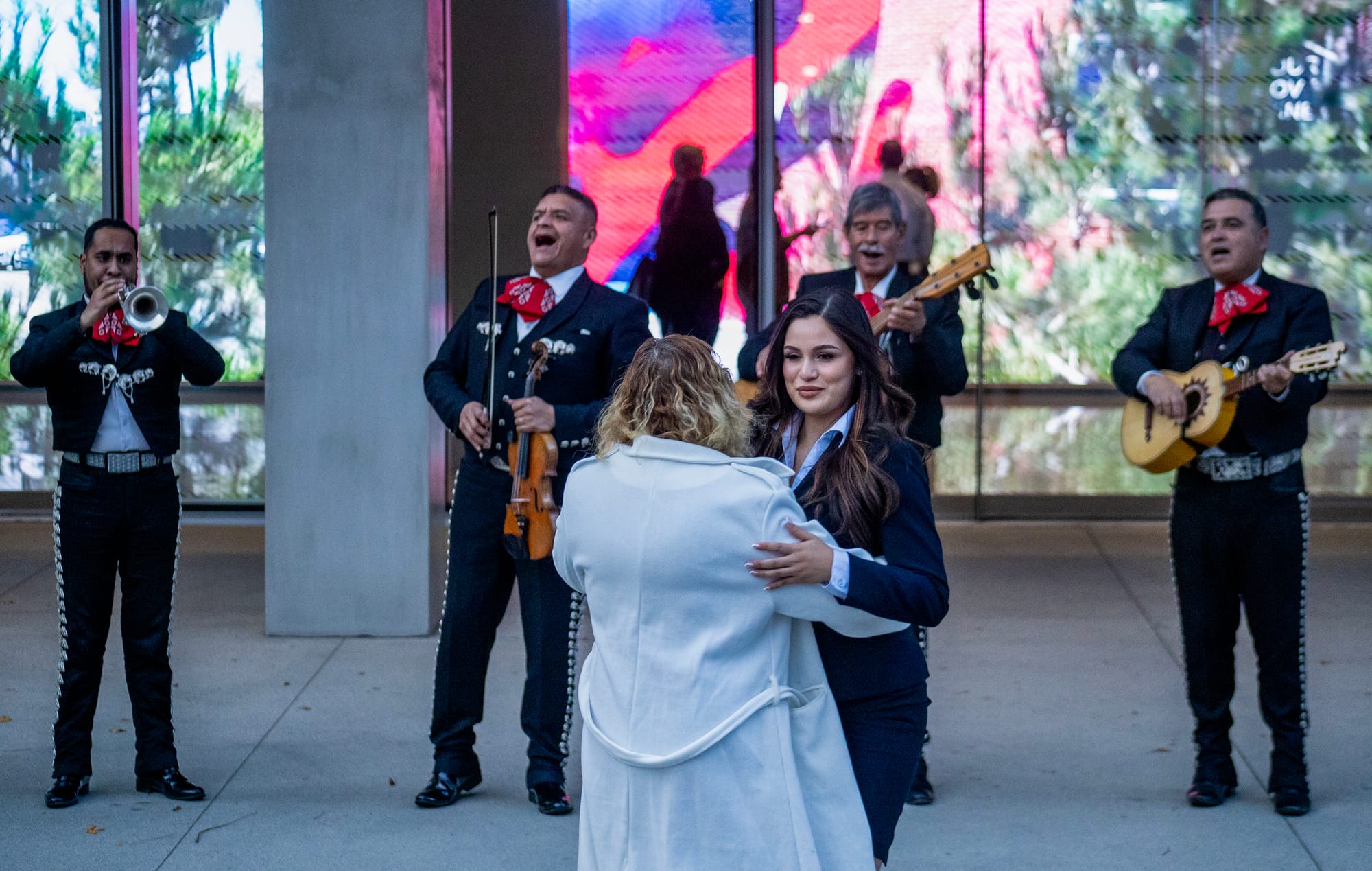 A young woman dances with another woman in a white coat with a mariachi band in the background.