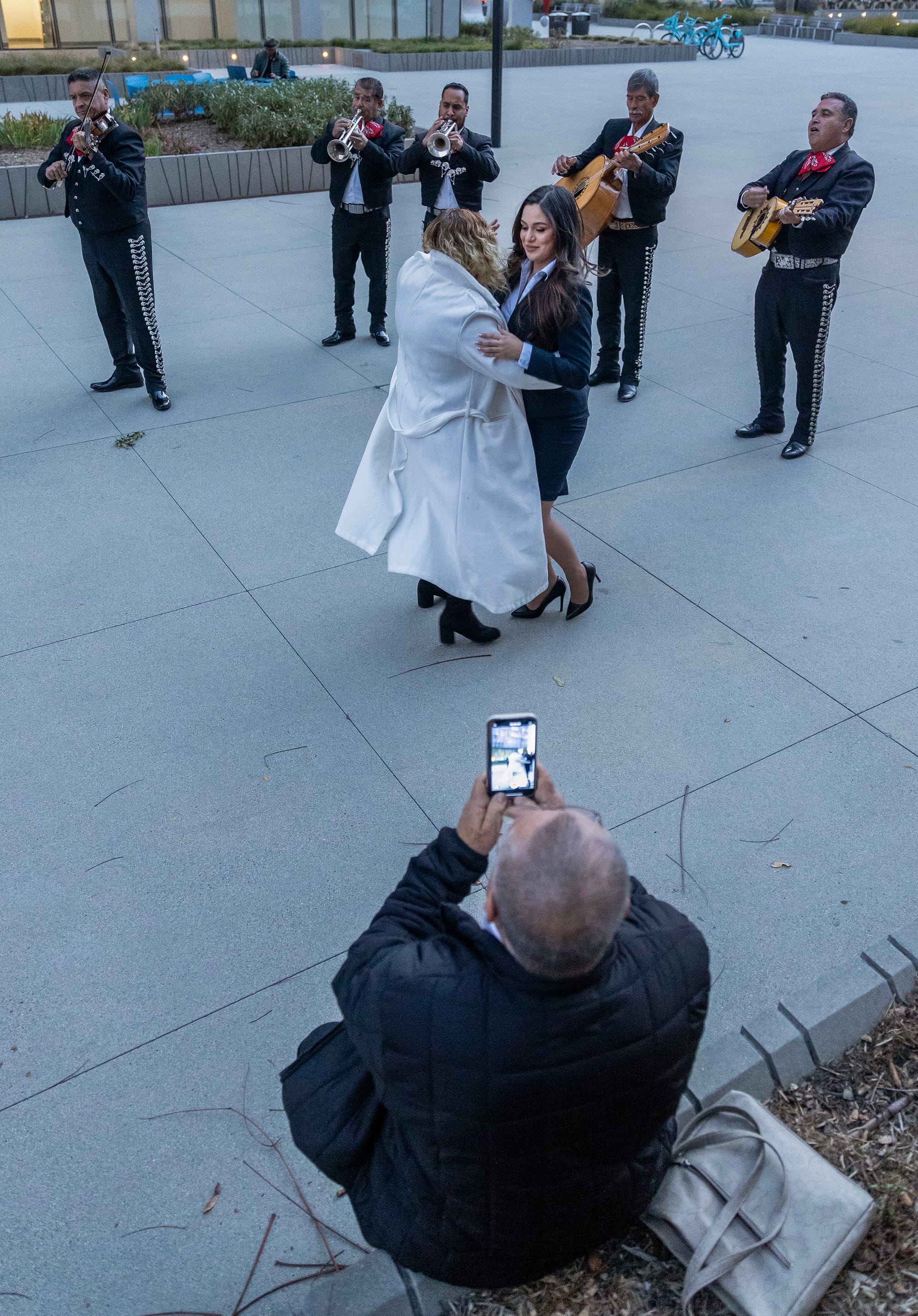 A man takes a video of a young woman dancing with another woman in a white coat with a mariachi band in the background.