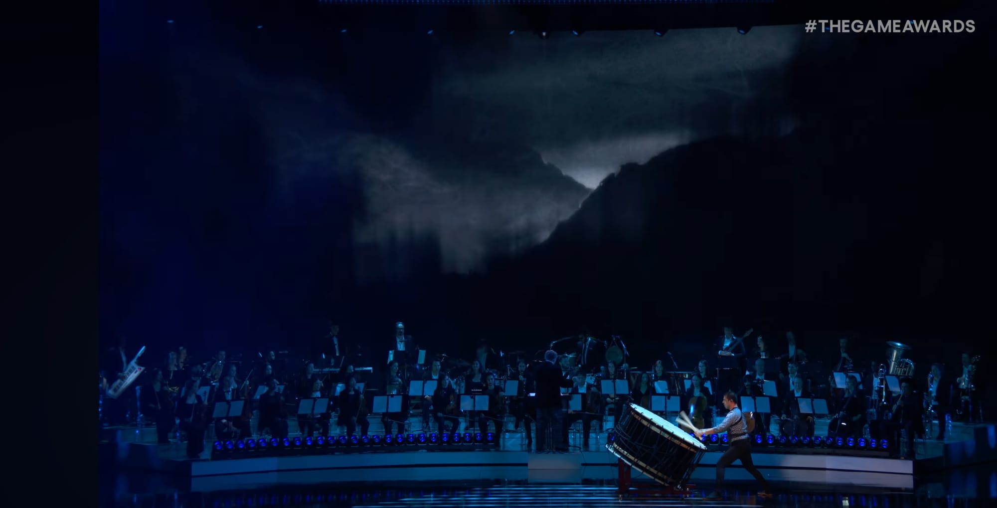 A man plays a large drum with an orchestra and a dark screen behind him.