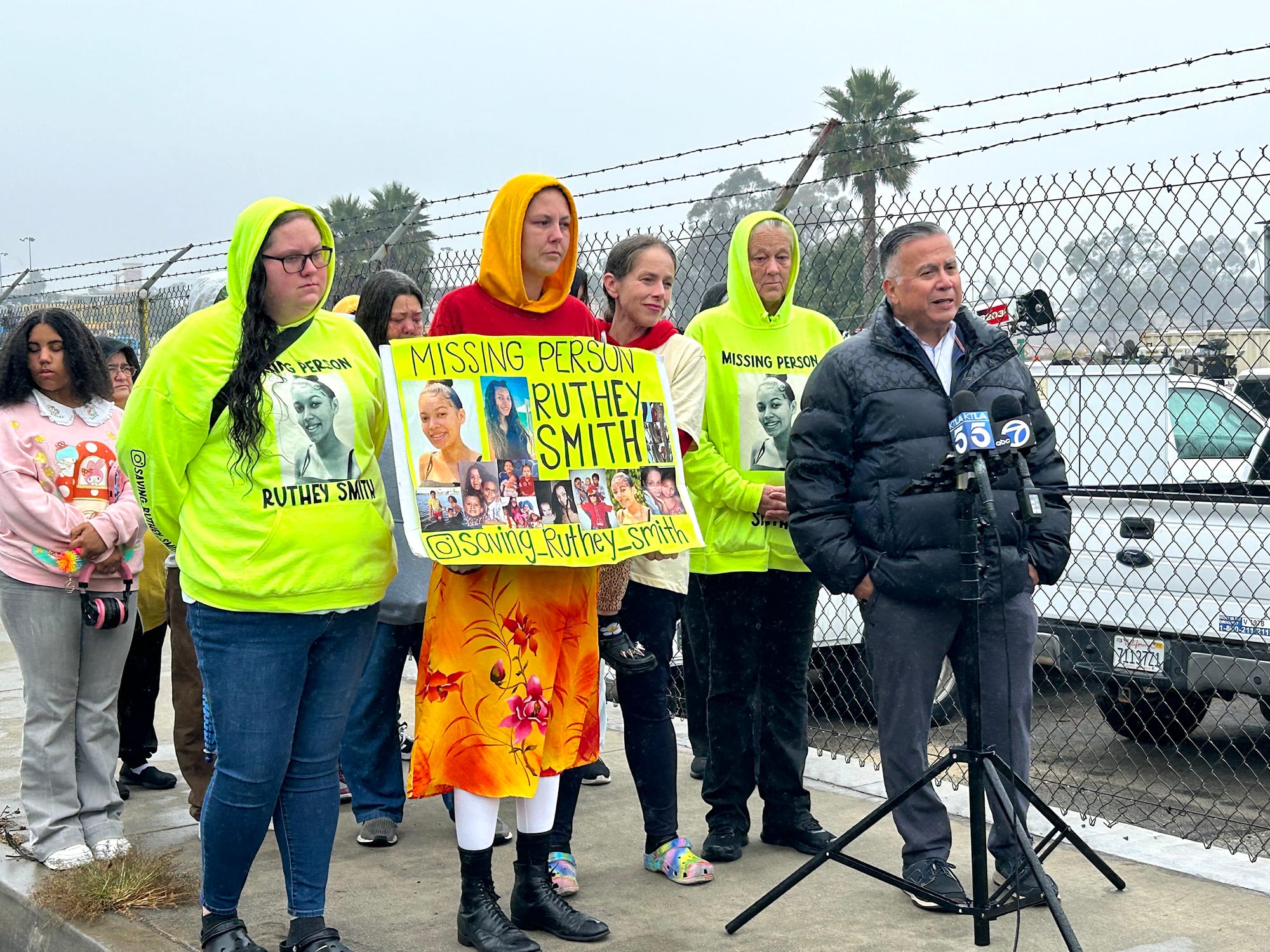 People stand in front of microphones next to a chain-link fence.