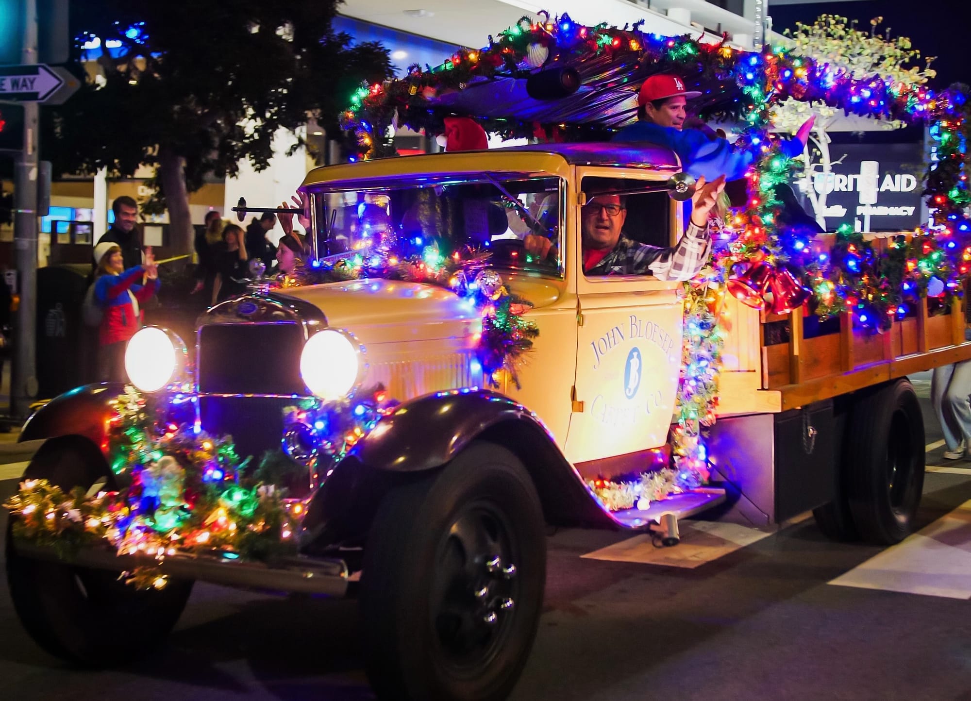 A yellow truck form the 1940s covered in bright lights rolls through a parade at night.