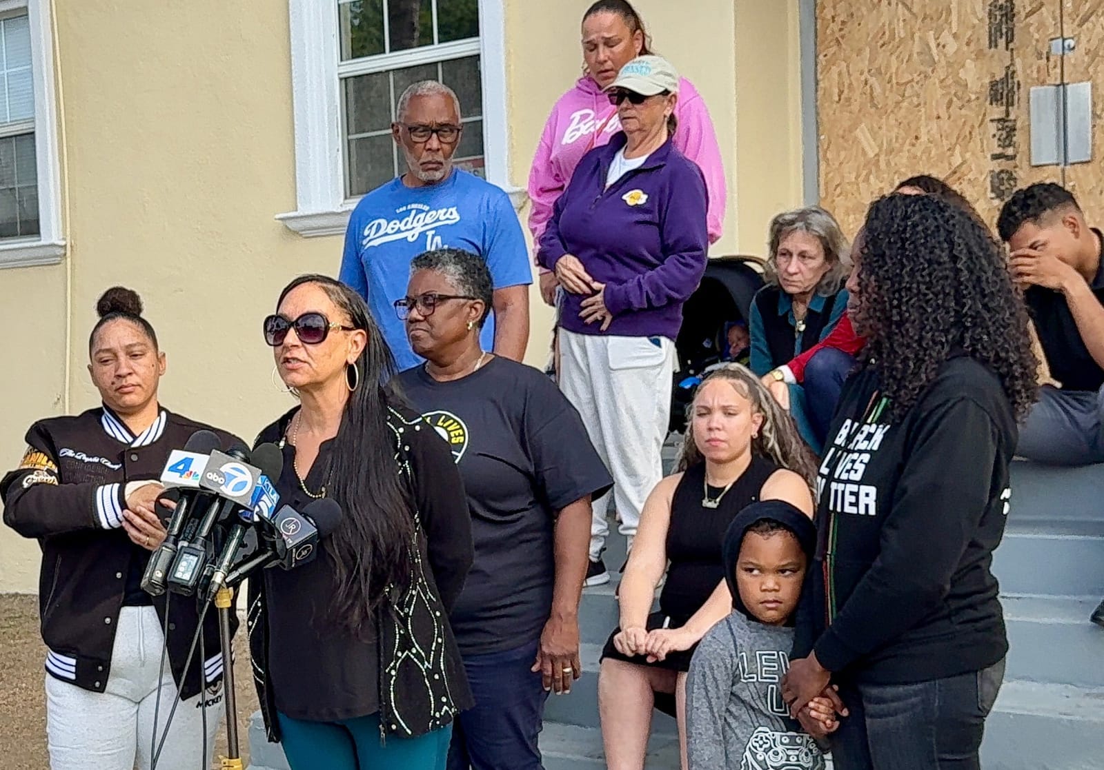 About a dozen people, including a small child, stand and sit on gray stairs in front of a group of microphones.