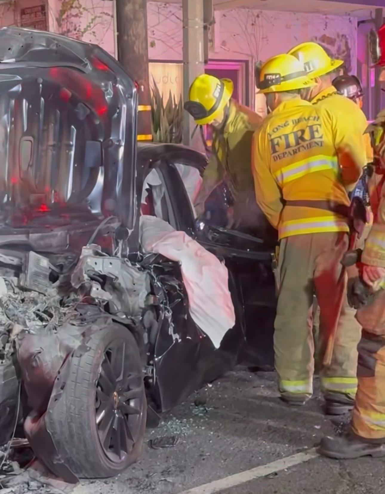 People in yellow jackets that say "Long Beach Fire Department" on the back stand next to a wrecked black car.