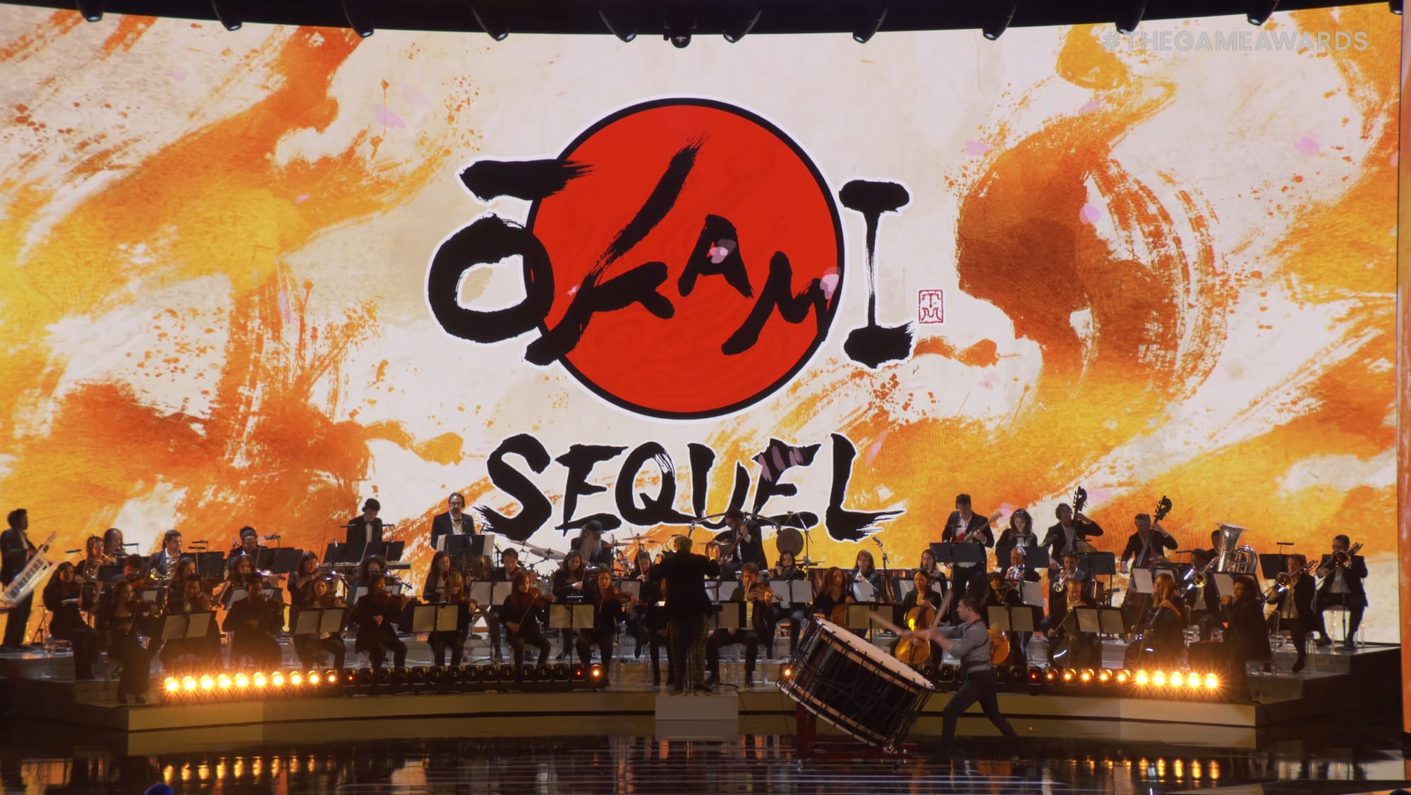 An orchestra sits on a stage with a bright white and orange backdrop that reads "Okami sequel."