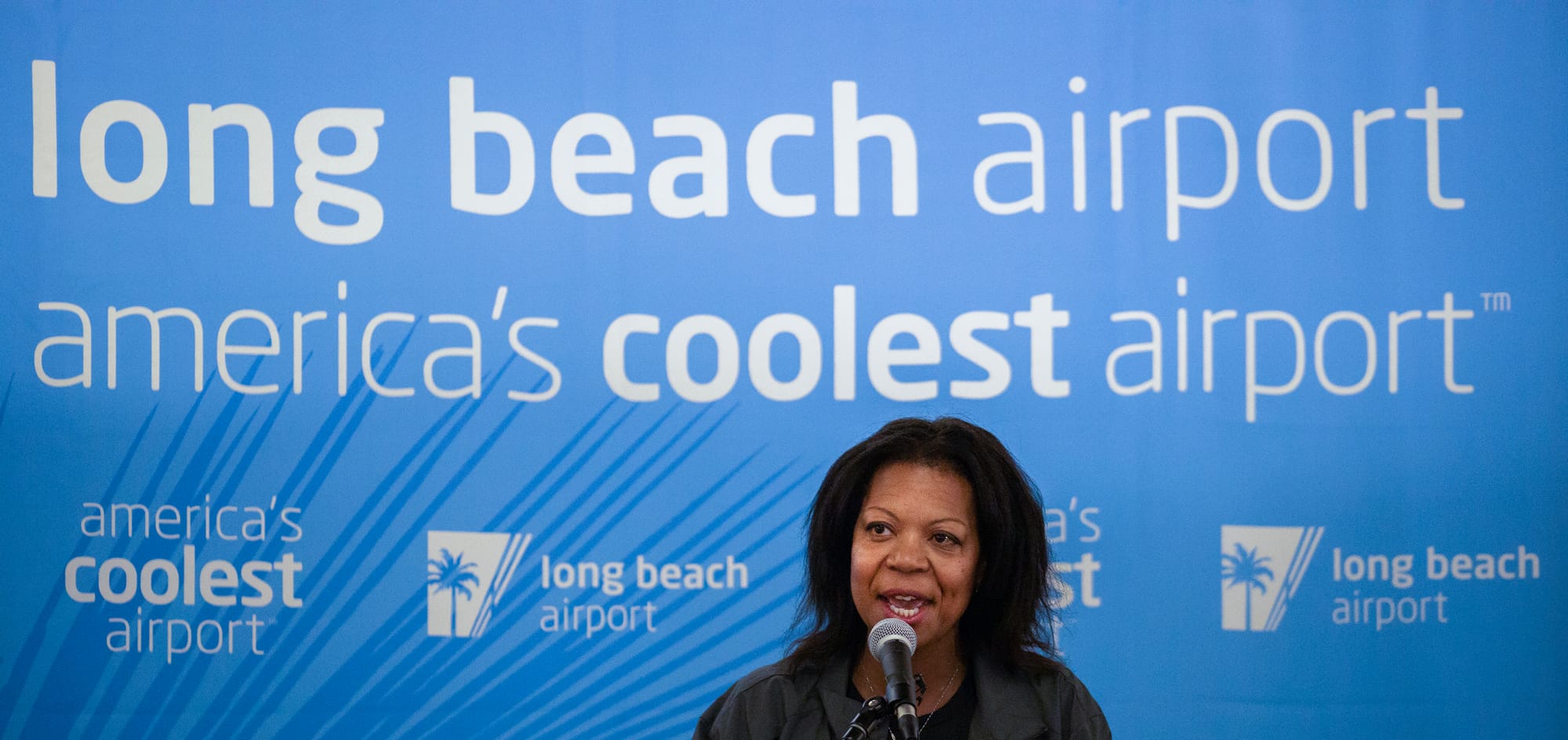 A Black woman speaks at a microphone in front of a blue banner that reads "Long Beach Airport" and "American's coolest airport."