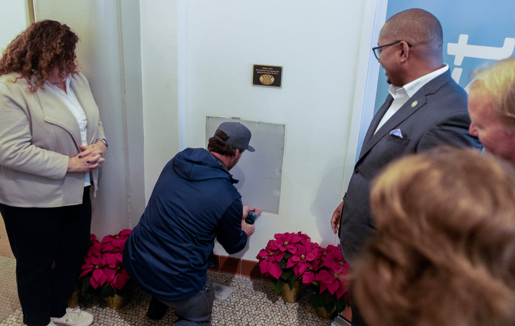 A man wearing a hoodie and a hat drills a panel into a wall as others watch.