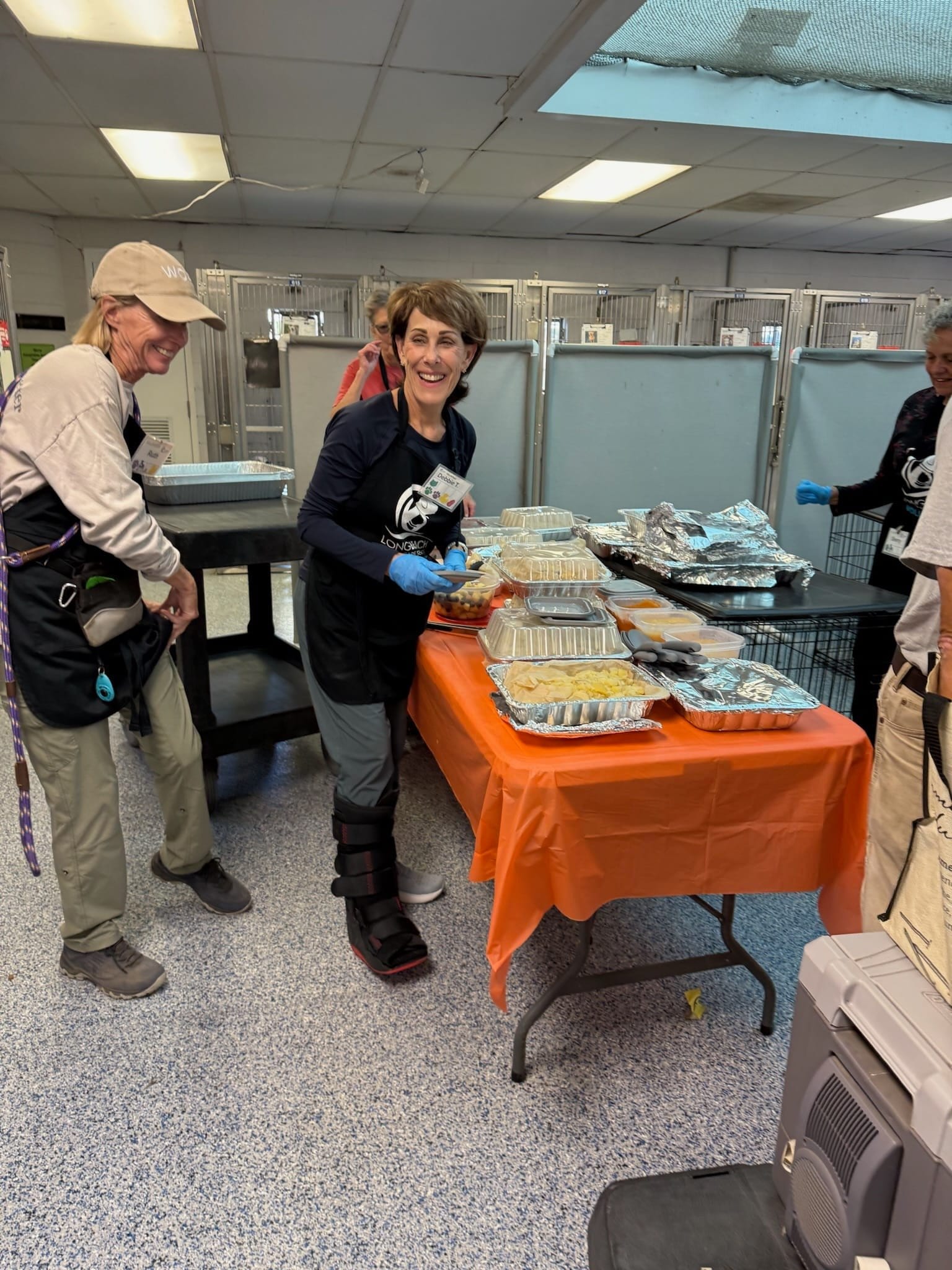 A woman smiles while standing next to a table covered in a orange fabric and topped with many trays of food.