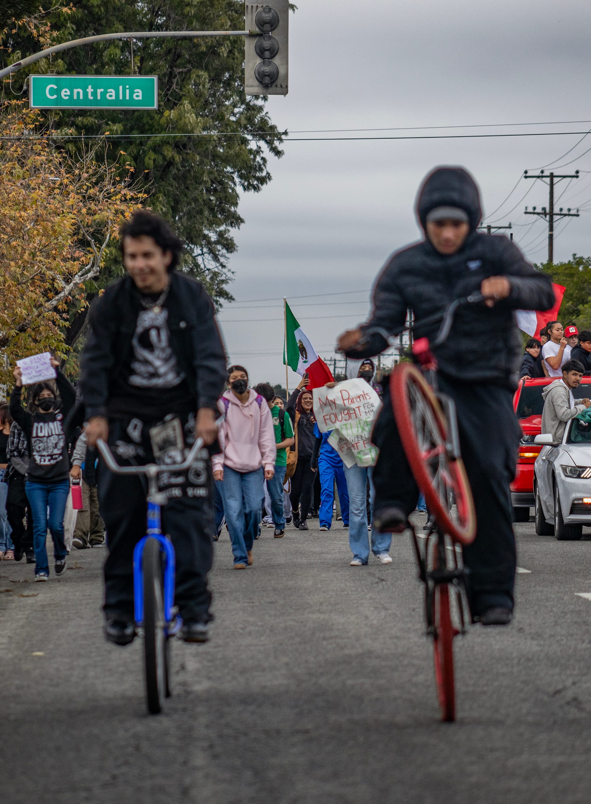 Two people ride bikes in front of other people marching in the street.