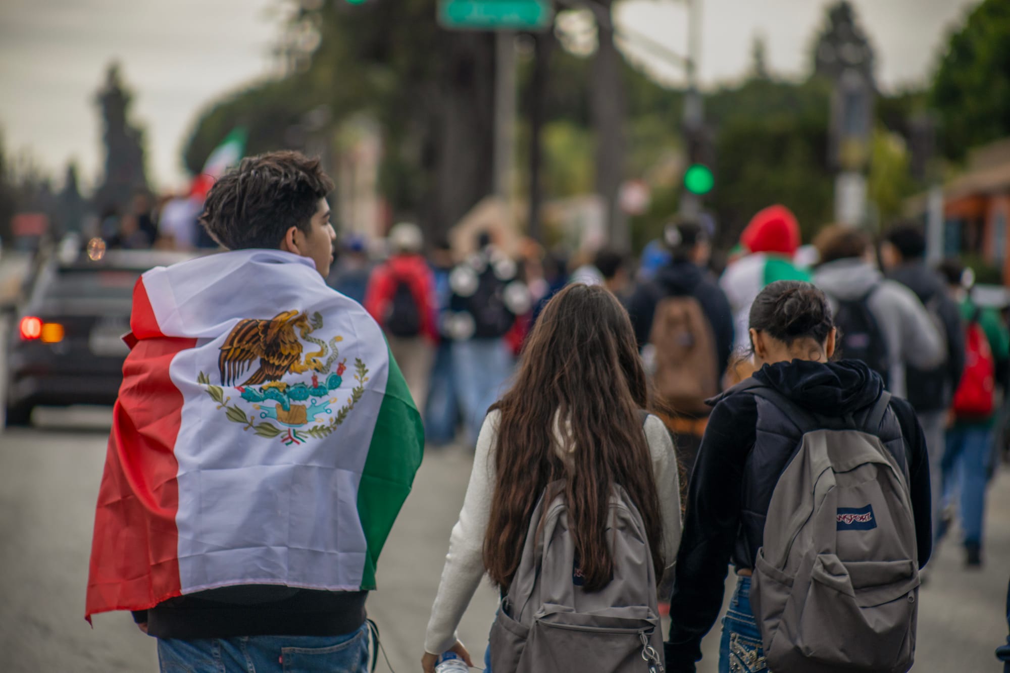 Three young people, one draped in the Mexican flag, walk down the street.