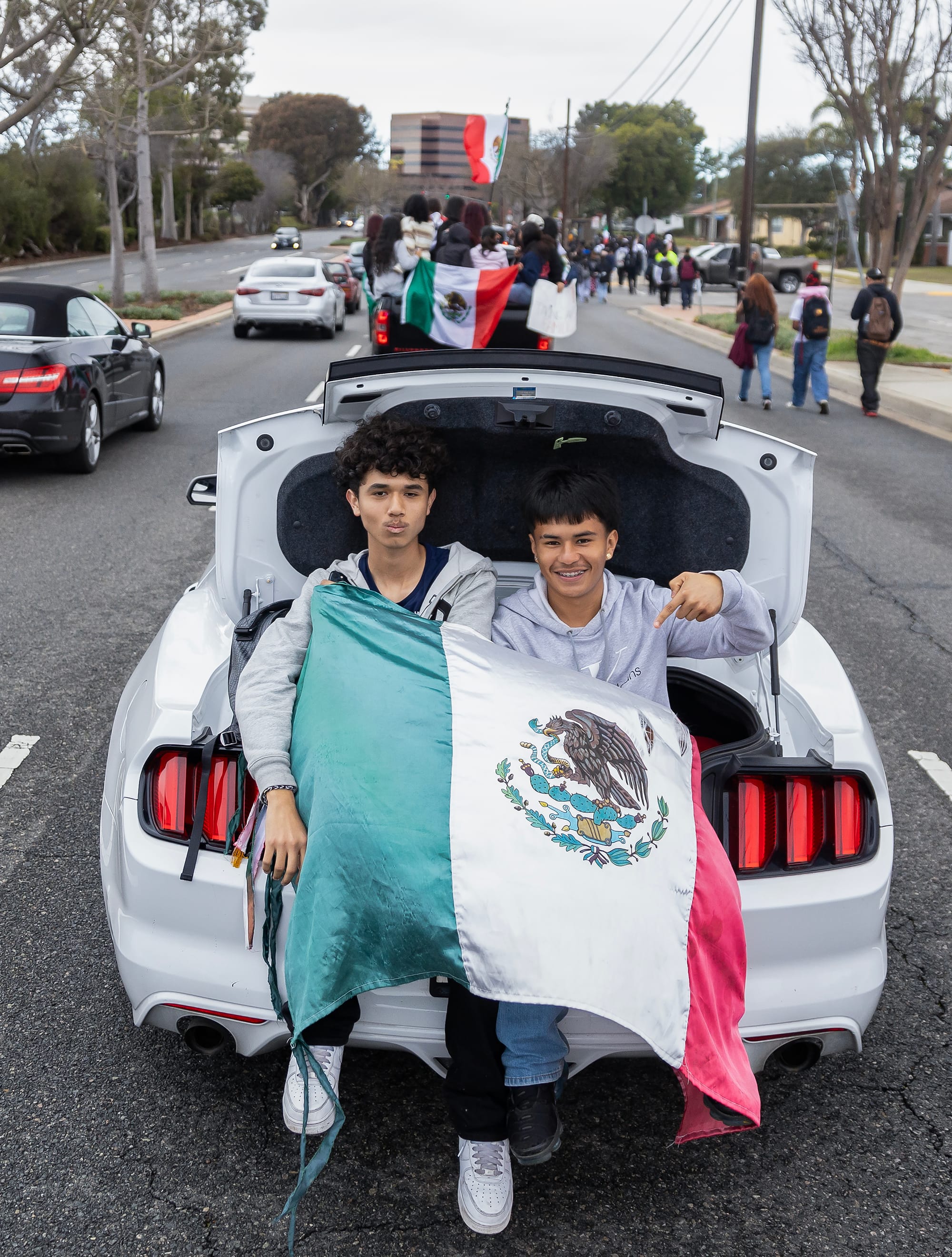 Two teens hold a Mexican flag as they sit in the trunk of a white car.