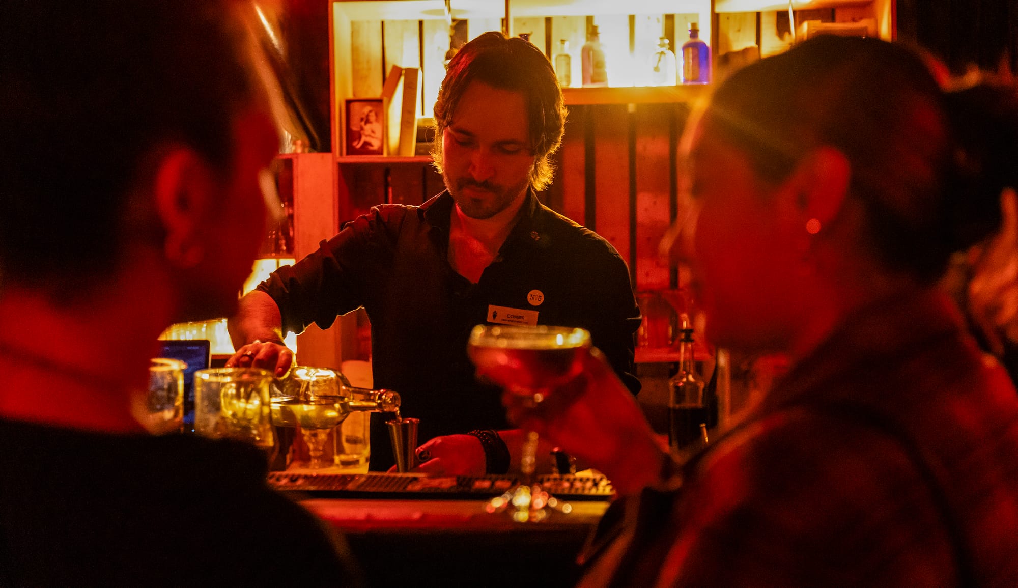 A man pours liquid into a measuring cup while a woman holds a cocktail glass.