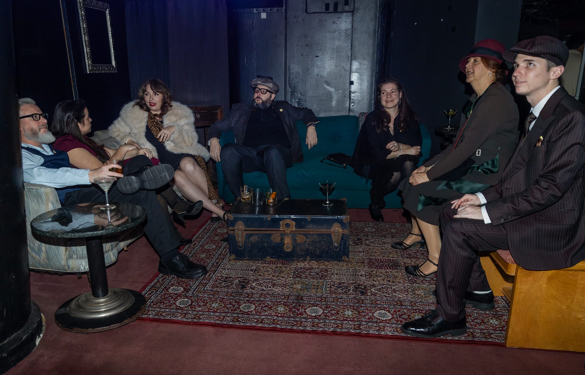 Seven people sit on couches next to a blue trunk.