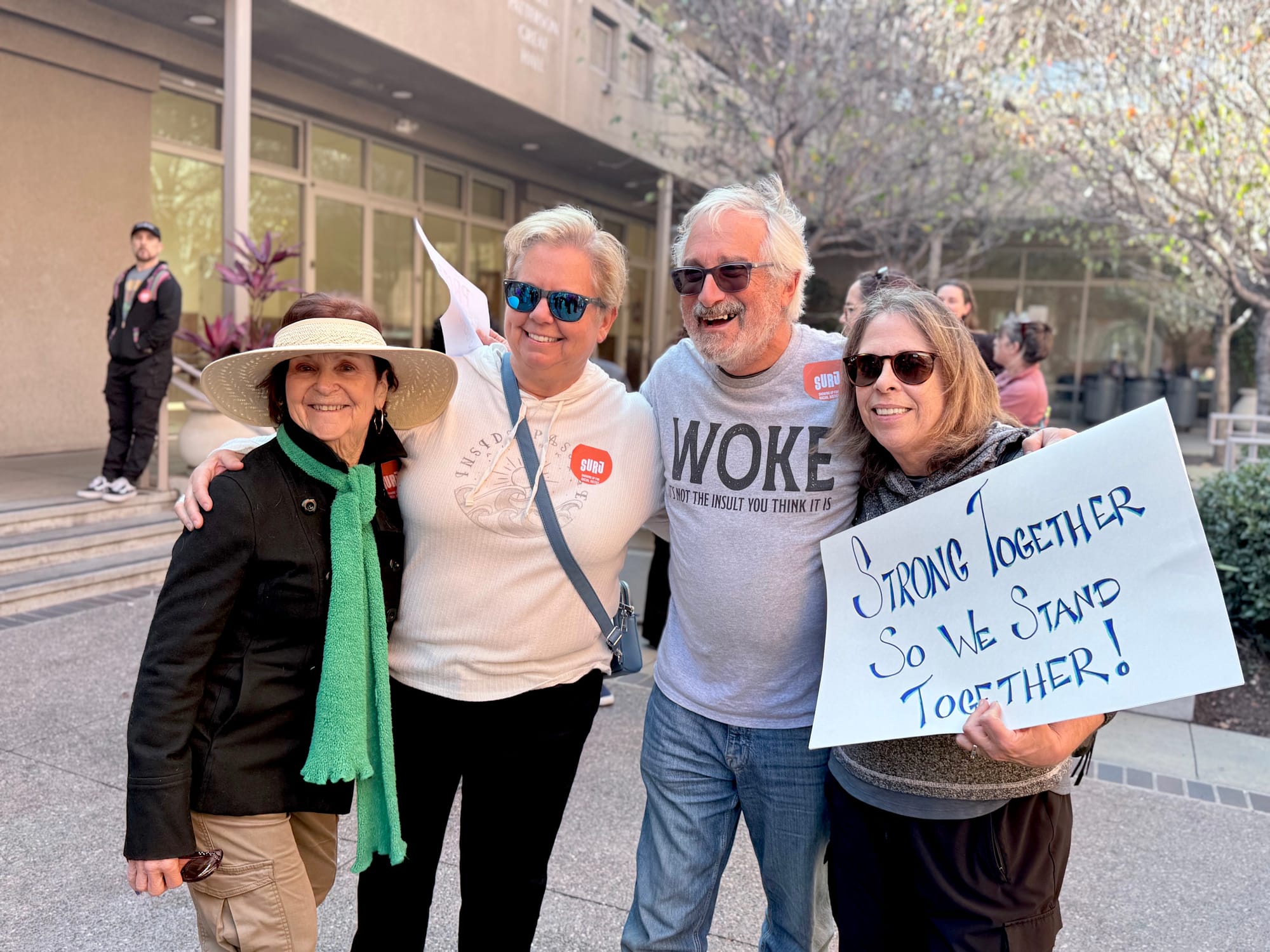 Four people smile and stand shoulder to shoulder next to a sign saying "Strong Together So We Stand Together!"