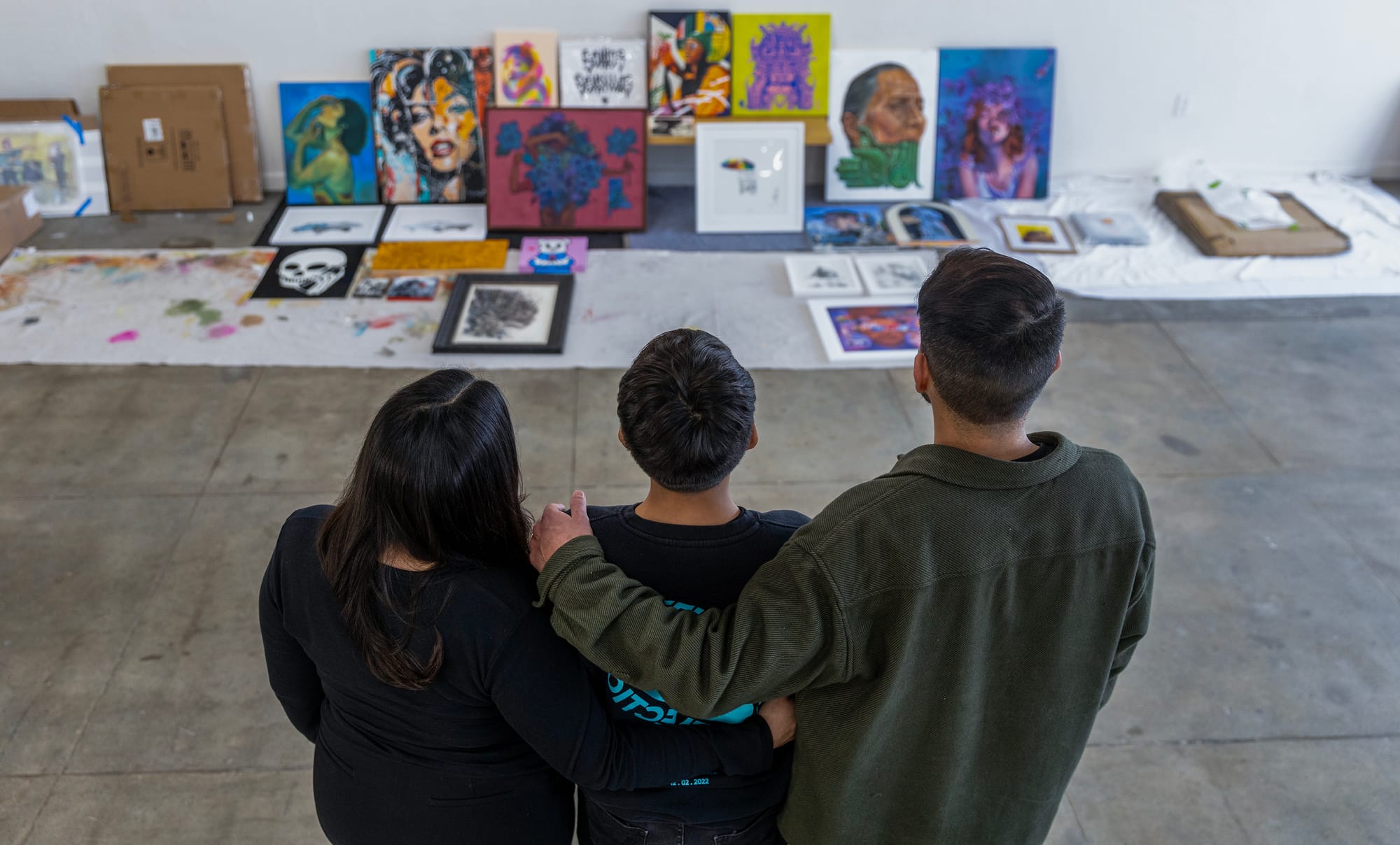 A man and woman stand with their arms around a teenage boy as they face away from the camera and look at colorful artwork.