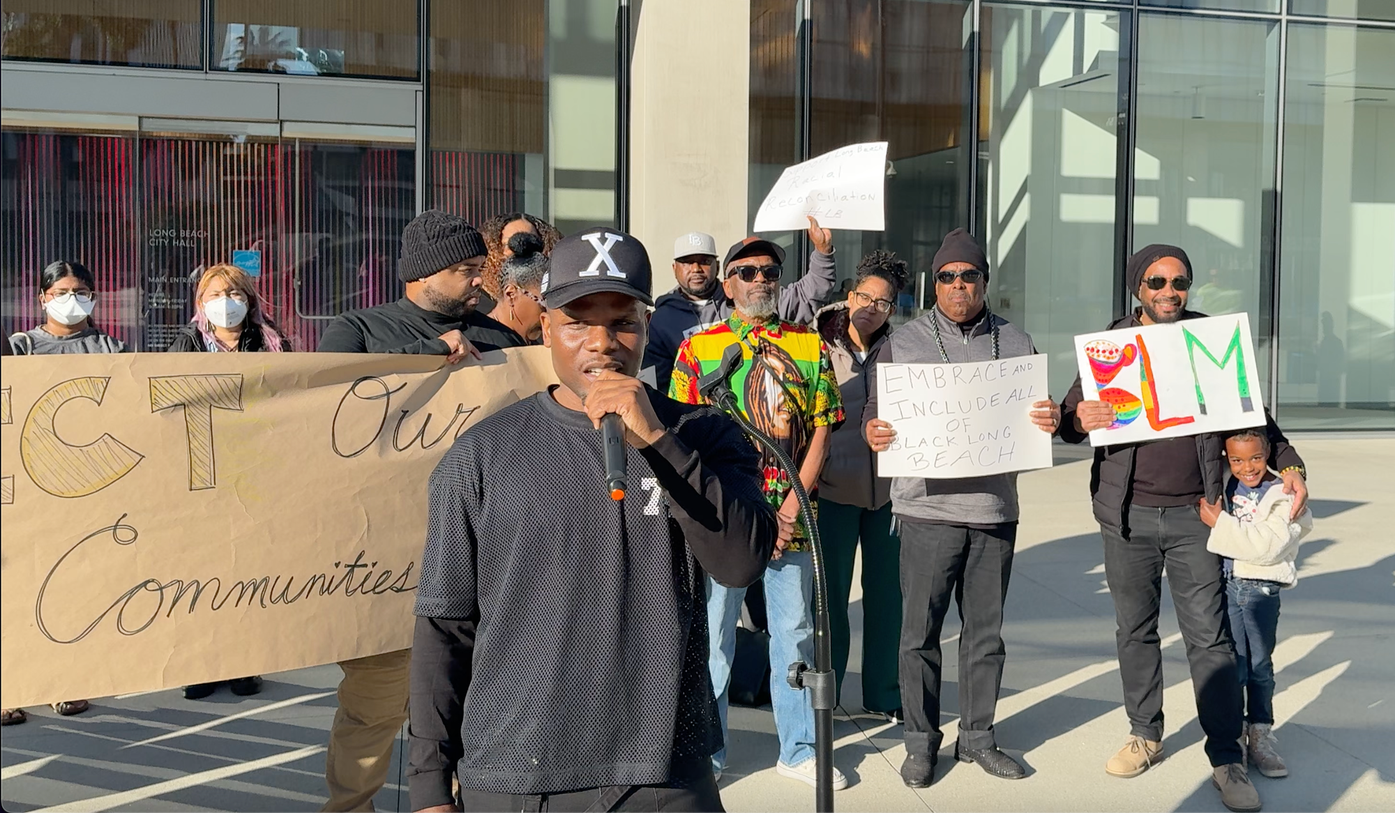 About a dozen people holding handmade signs stand in front of a glassy office building.