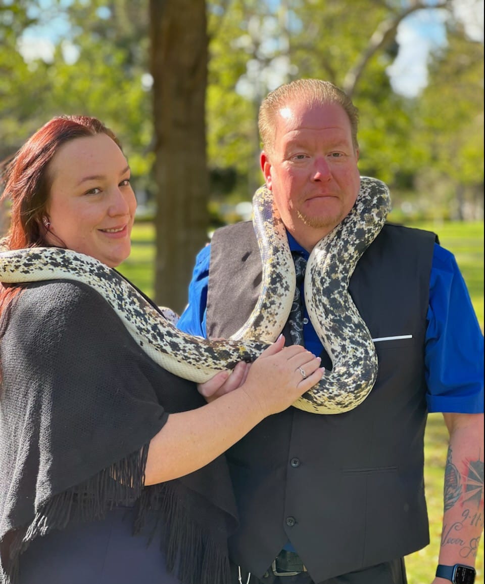 A man and a woman pose with a large snake draped over their shoulders.