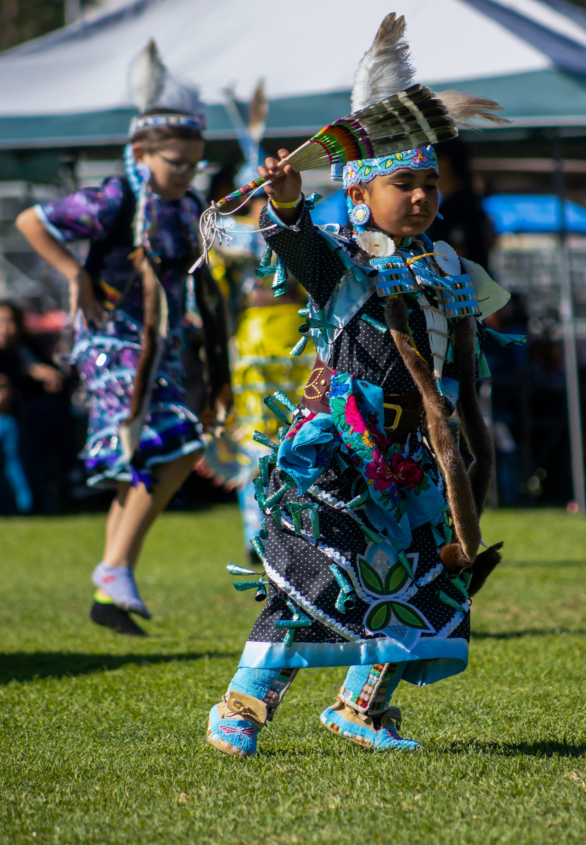 Children in colorful Native American garb dance outside on the grass.