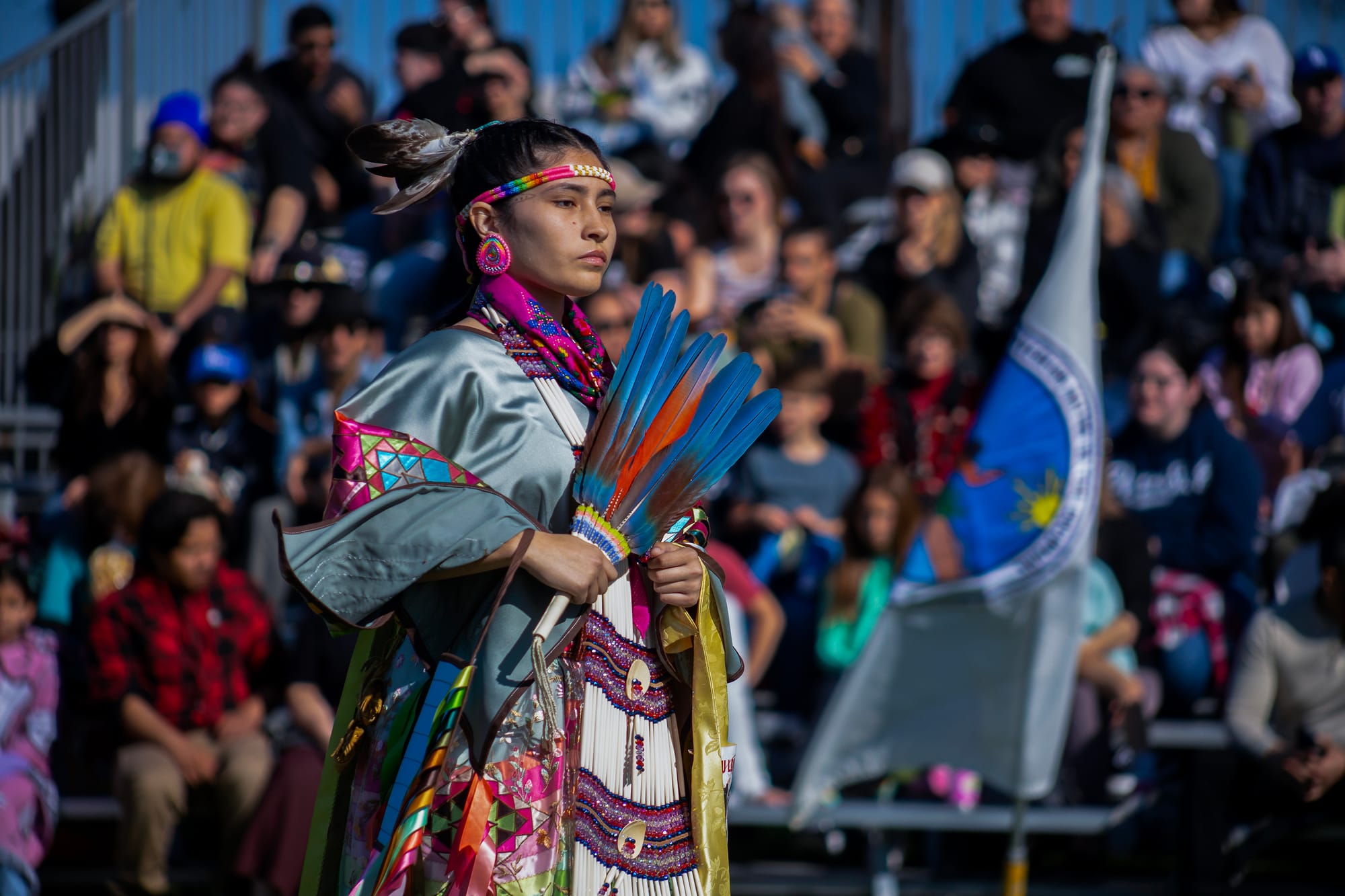 A woman in colorful Native American garb dances outside in front of a large group of people.