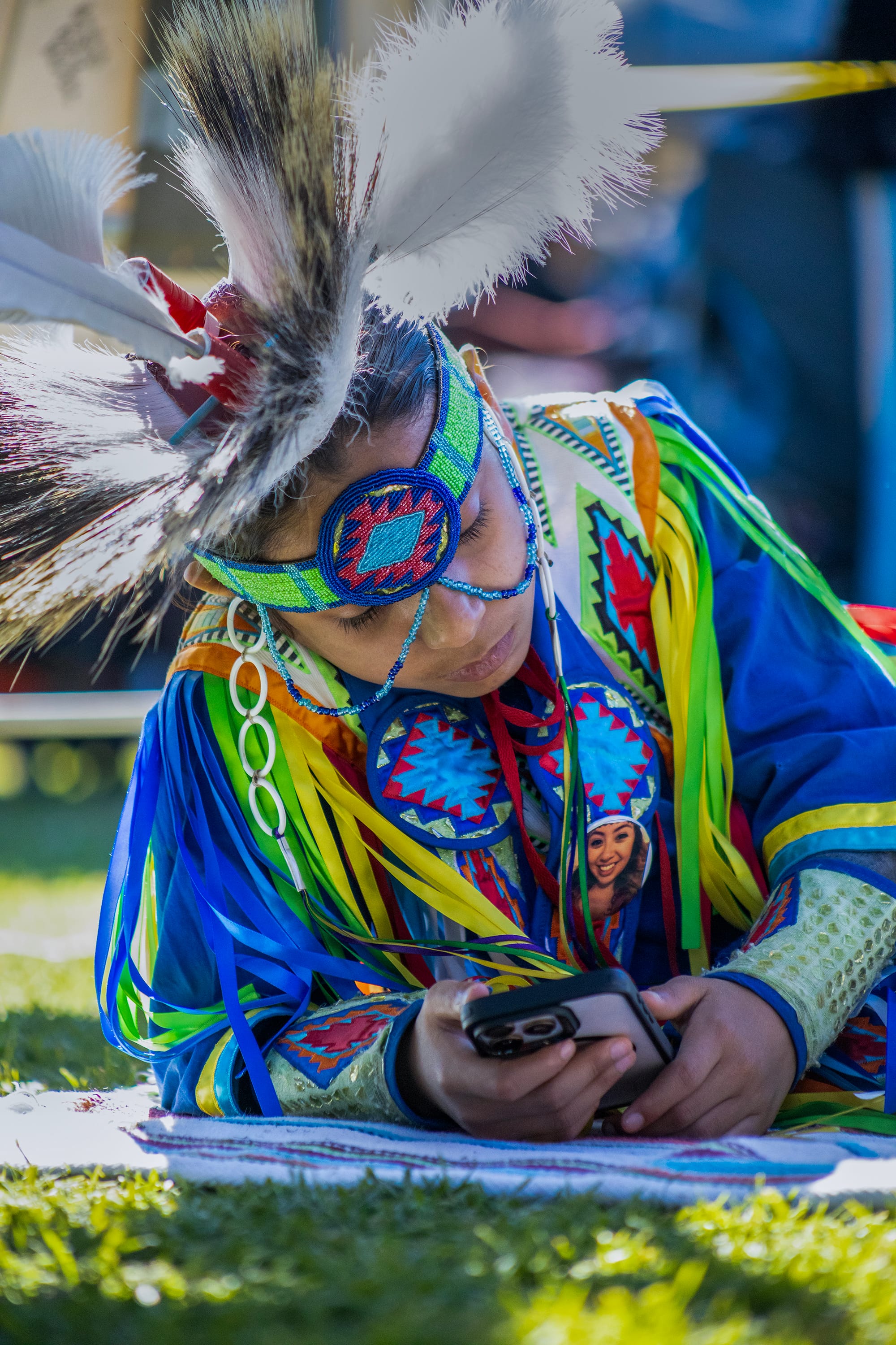 A boy in colorful Native American garb lies on the grass while checking his phone.