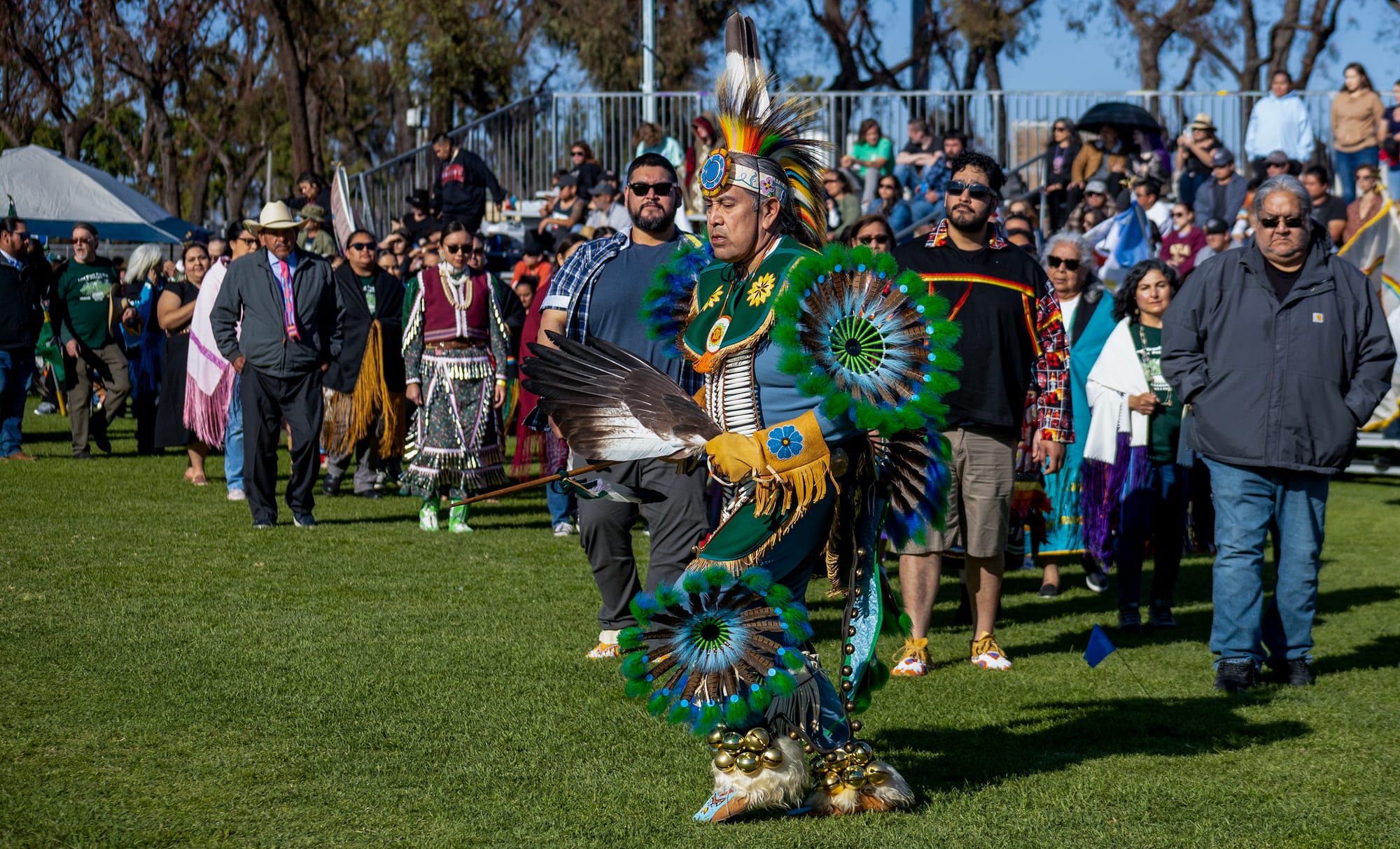 A person in colorful Native American garb dances outside in front of a large group of people.