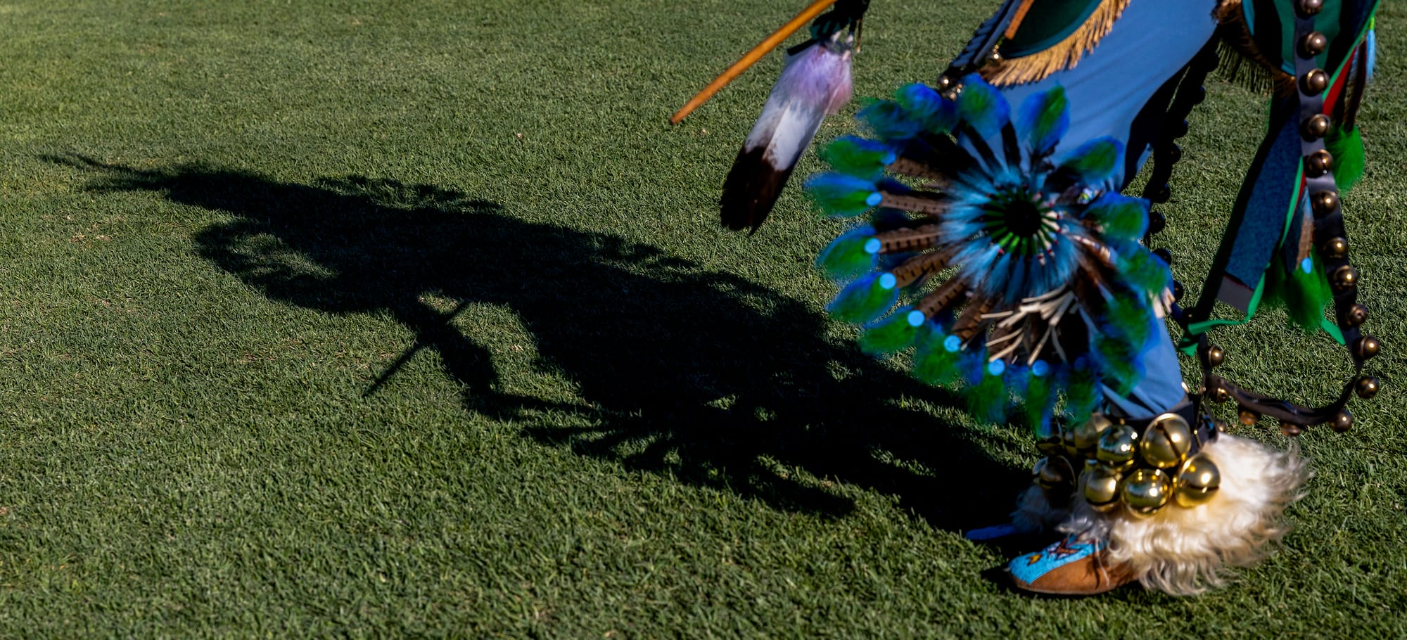 The shadow cast on a lawn from a man dancing in Native American garb.