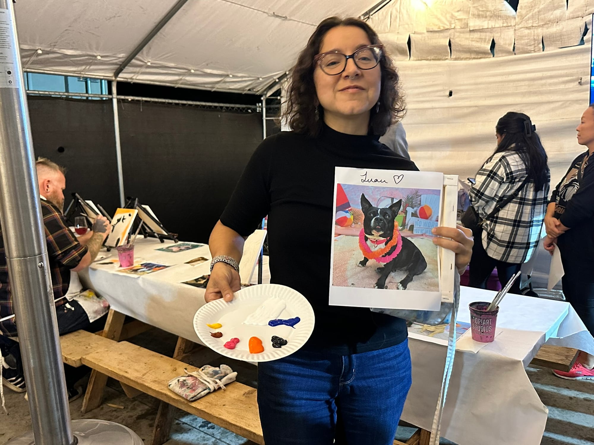 A woman holds a portrait of a dog and a paper plate covered in paint.