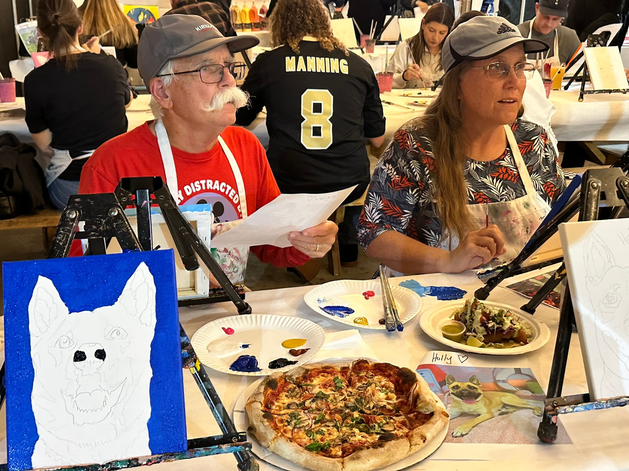 Two people sit at a table laden with pizza and painting supplies.
