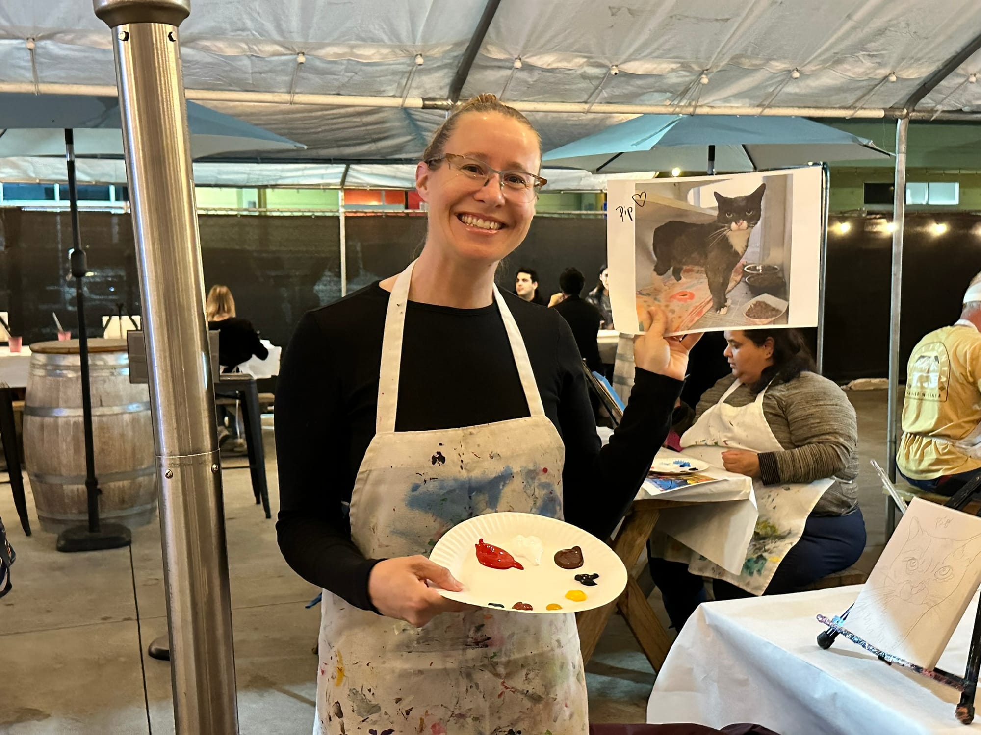 A woman holds a painting of a cat and a paper plate covered in paint.